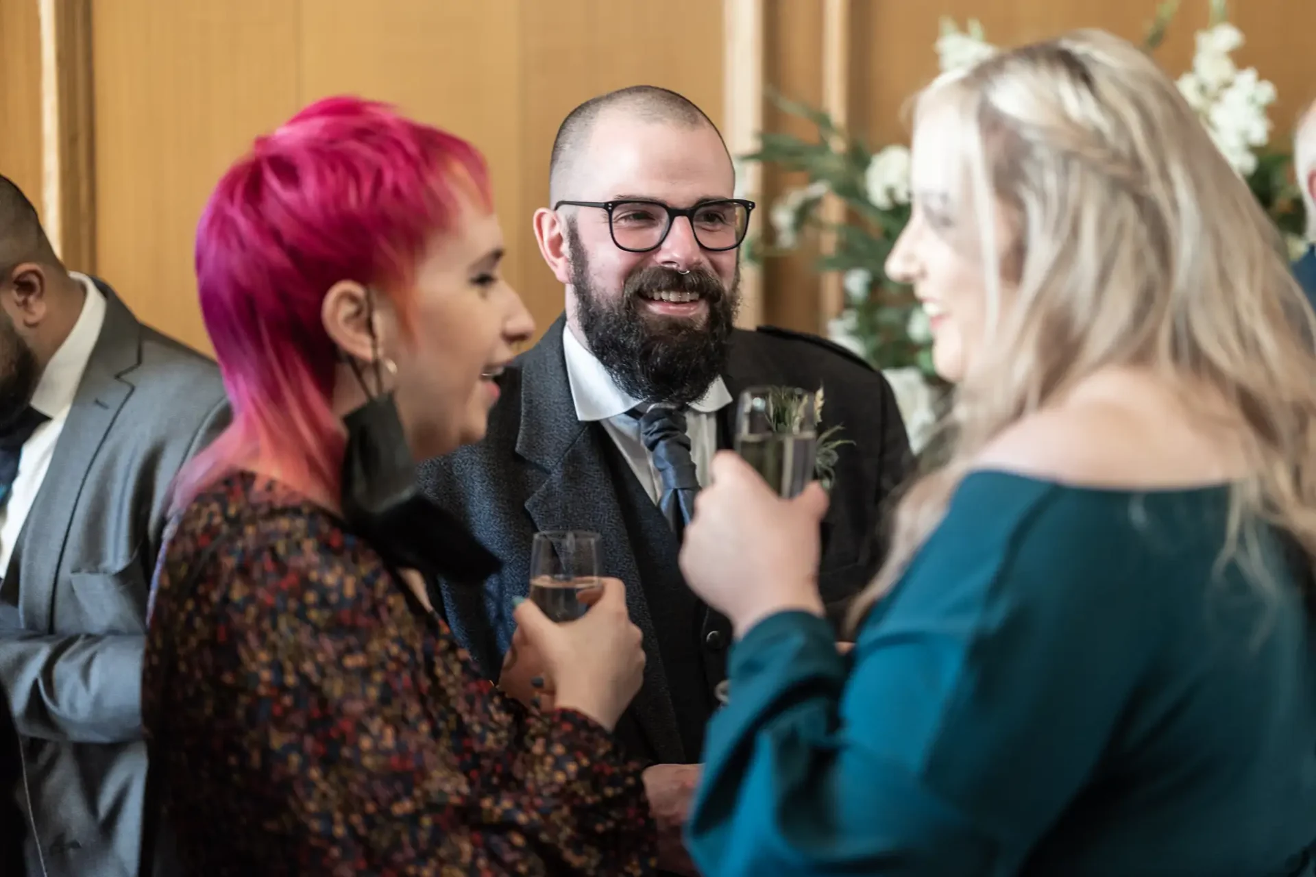 Group of people conversing happily at a formal event, one man in a suit and two women, one with pink hair, holding drinks.
