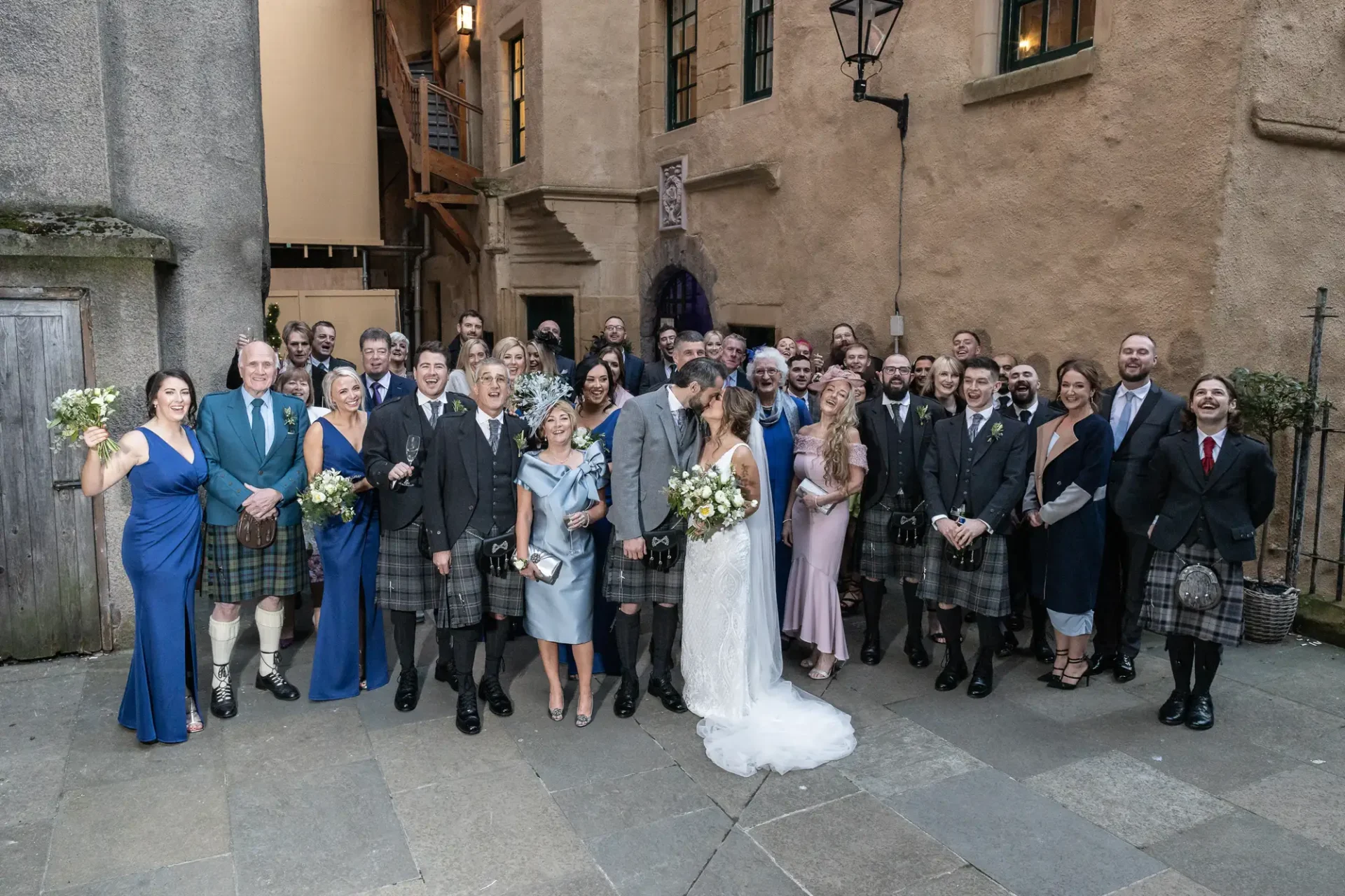 A large wedding group photo featuring a bride and groom kissing, surrounded by guests in formal attire, many in scottish kilts, in a historic courtyard.