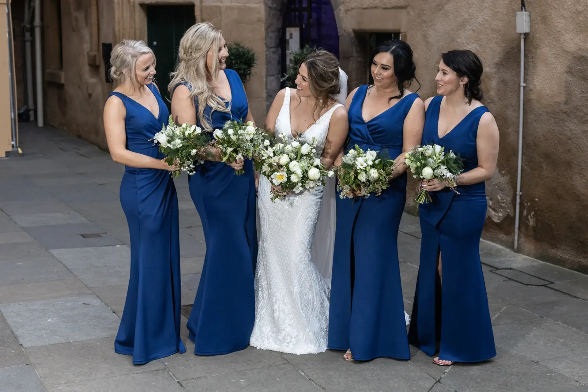 Bride in a white dress surrounded by four bridesmaids in blue dresses, all holding bouquets, on a cobblestone street.