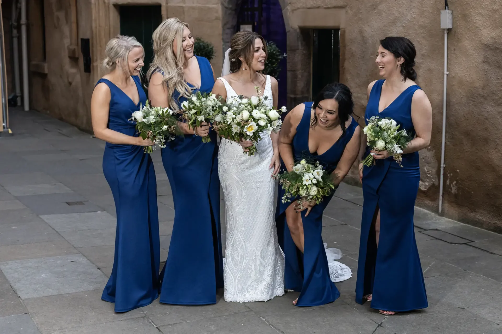 A bride and her bridesmaids, all in blue dresses and holding bouquets, laugh together on a city street.