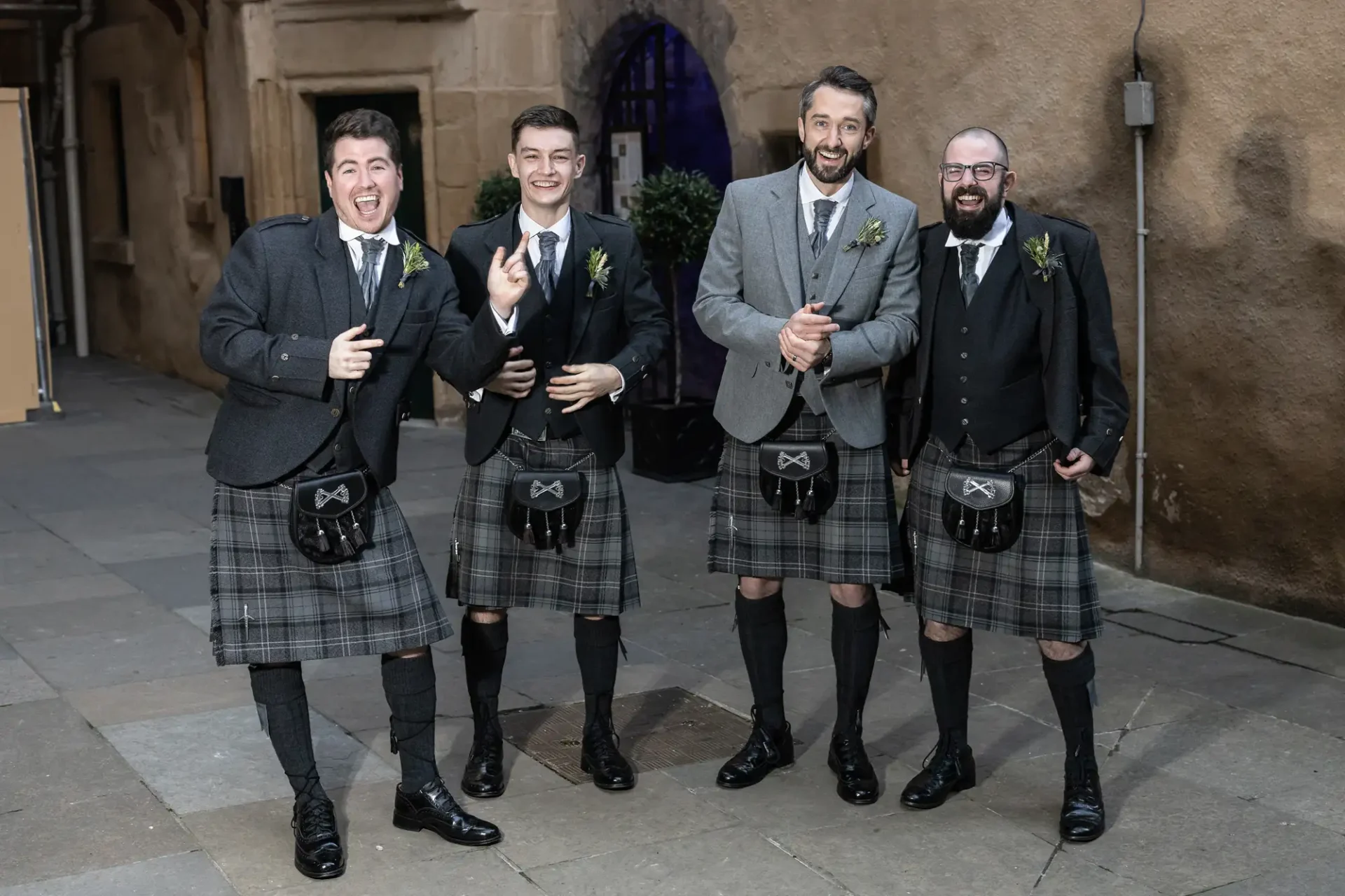 Four men in traditional scottish kilts and jackets, laughing and posing together at an outdoor event.