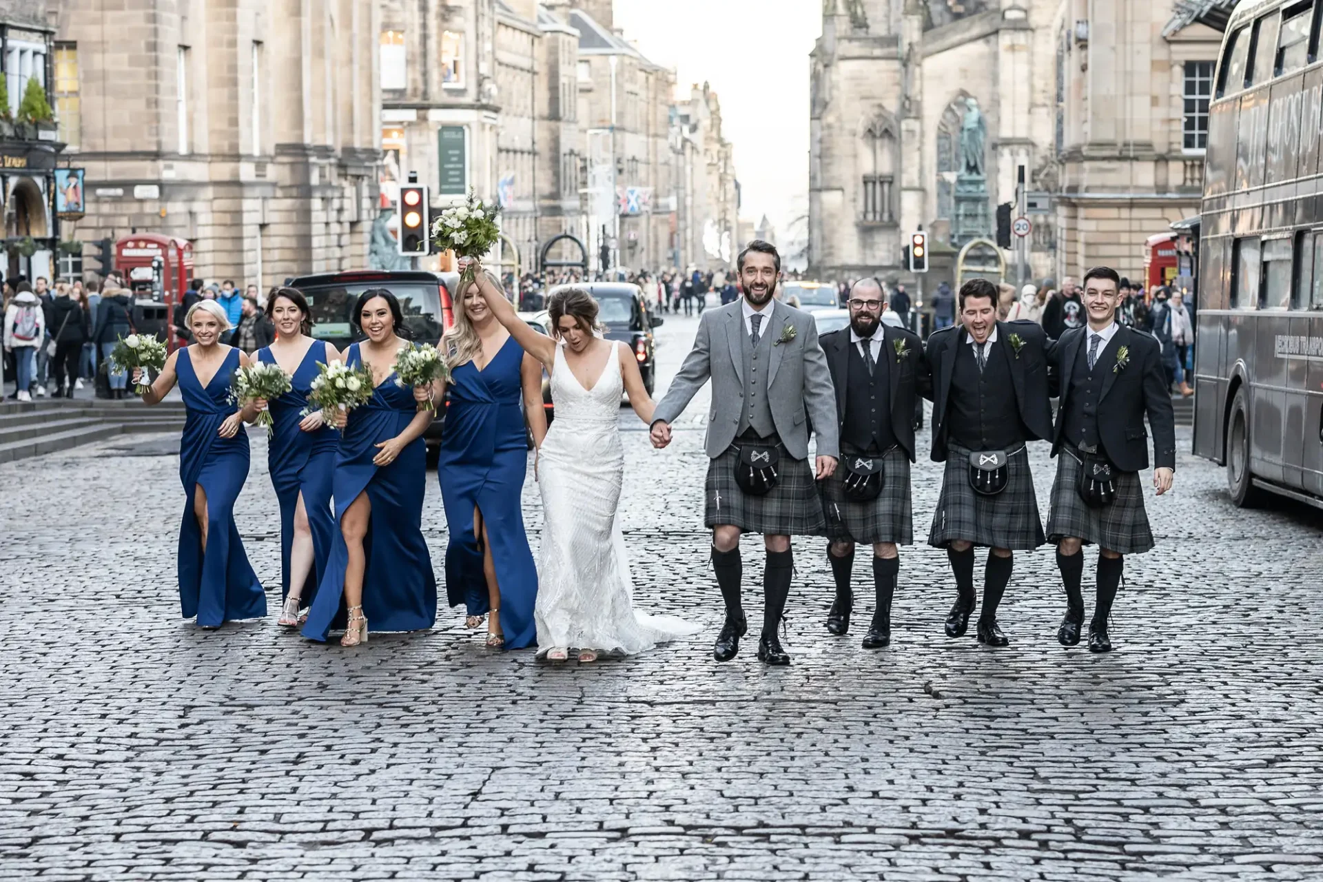 A wedding party, consisting of a bride, groom, and bridesmaids in blue dresses, along with groomsmen in kilts, joyfully walks down a cobblestone street in a city.