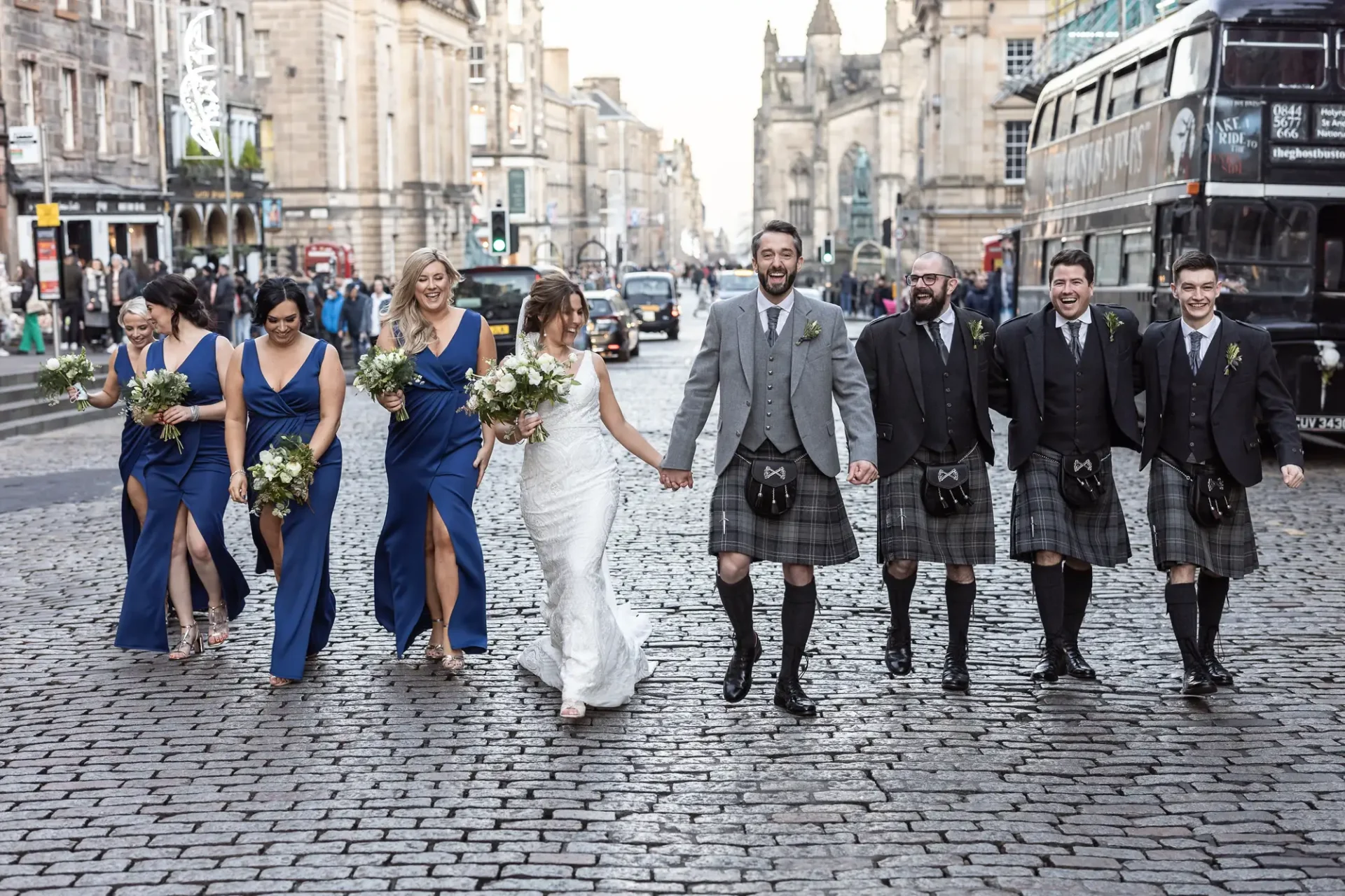 A bride and groom smiling and walking down a cobblestone street with their bridal party, men in kilts and women in blue dresses, in an urban setting.