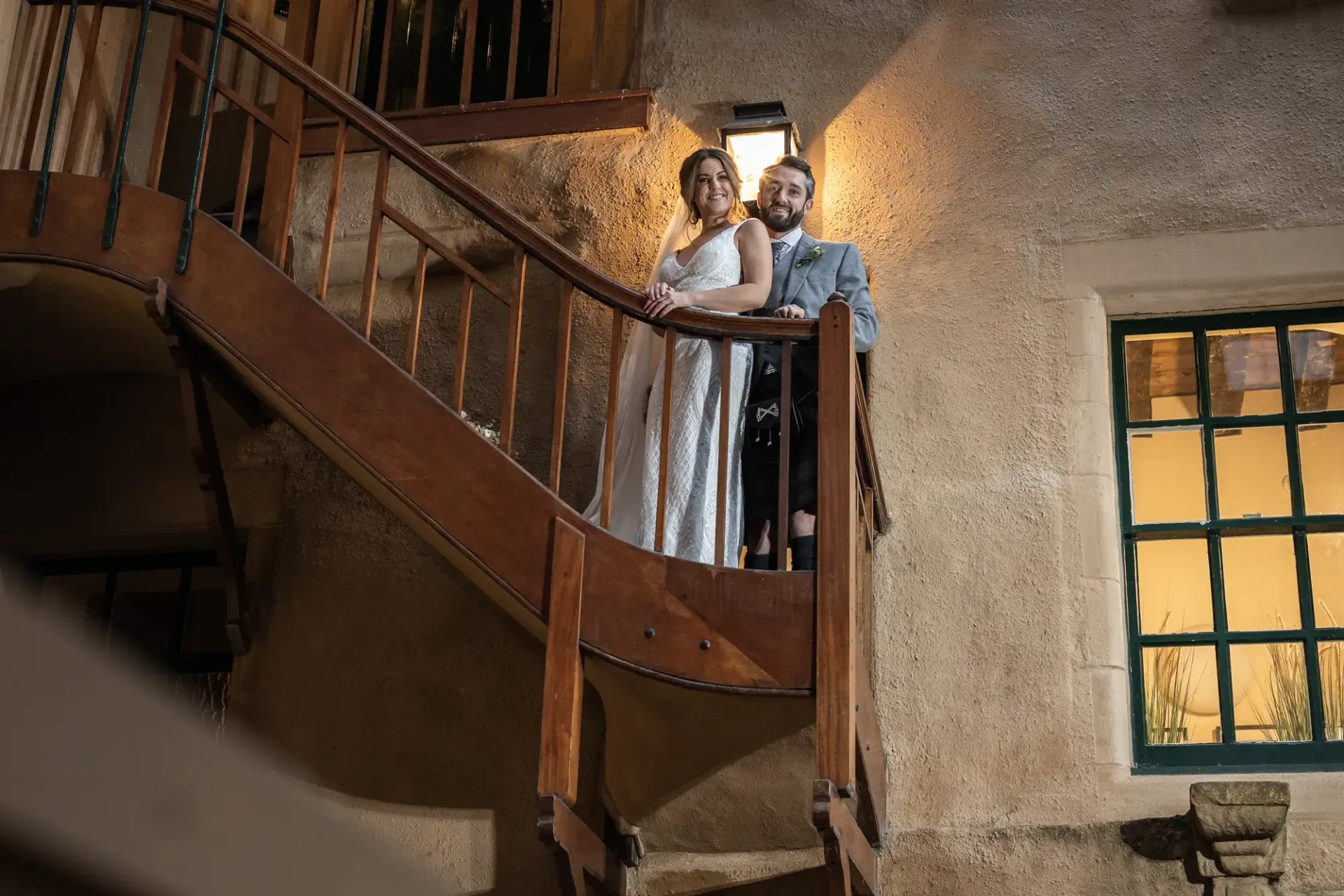 A bride and groom smiling, standing together on a curved wooden staircase in an elegant, warmly lit room.