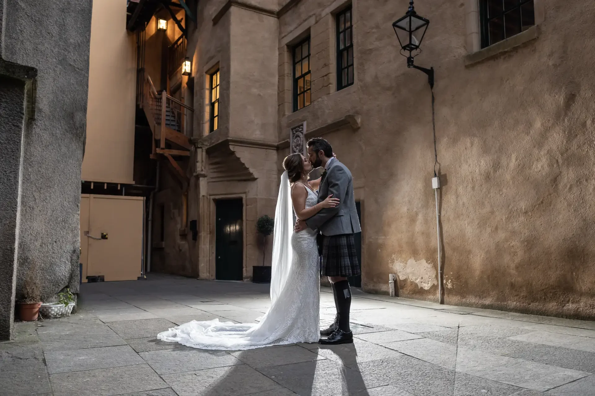 Bride and groom kissing in a sunlit alley, the bride in a long white dress and the groom in a tartan kilt.