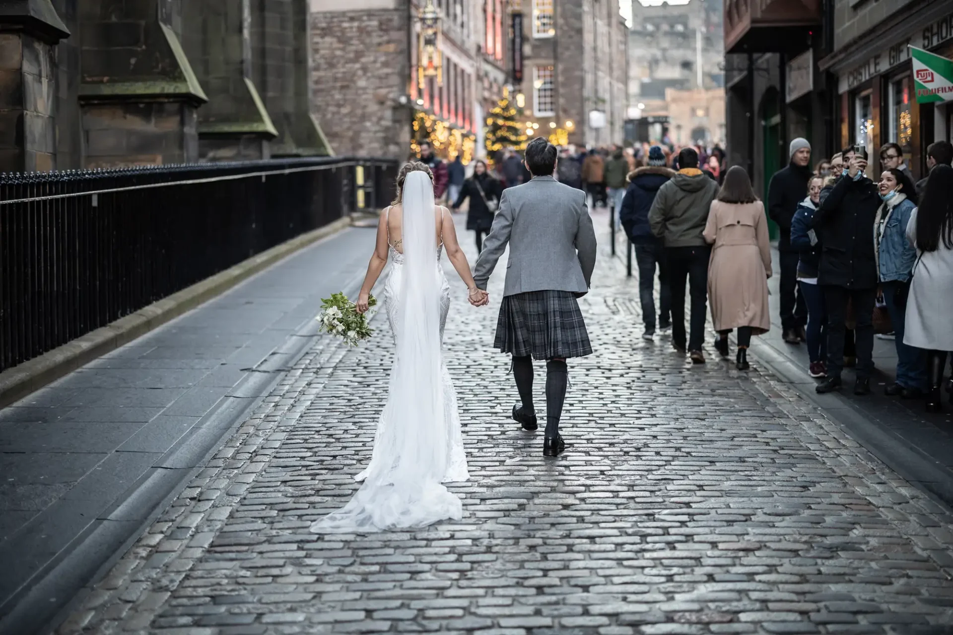 A bride in a white gown and a groom in a kilt holding hands, walking down a cobblestone street lined with pedestrians.