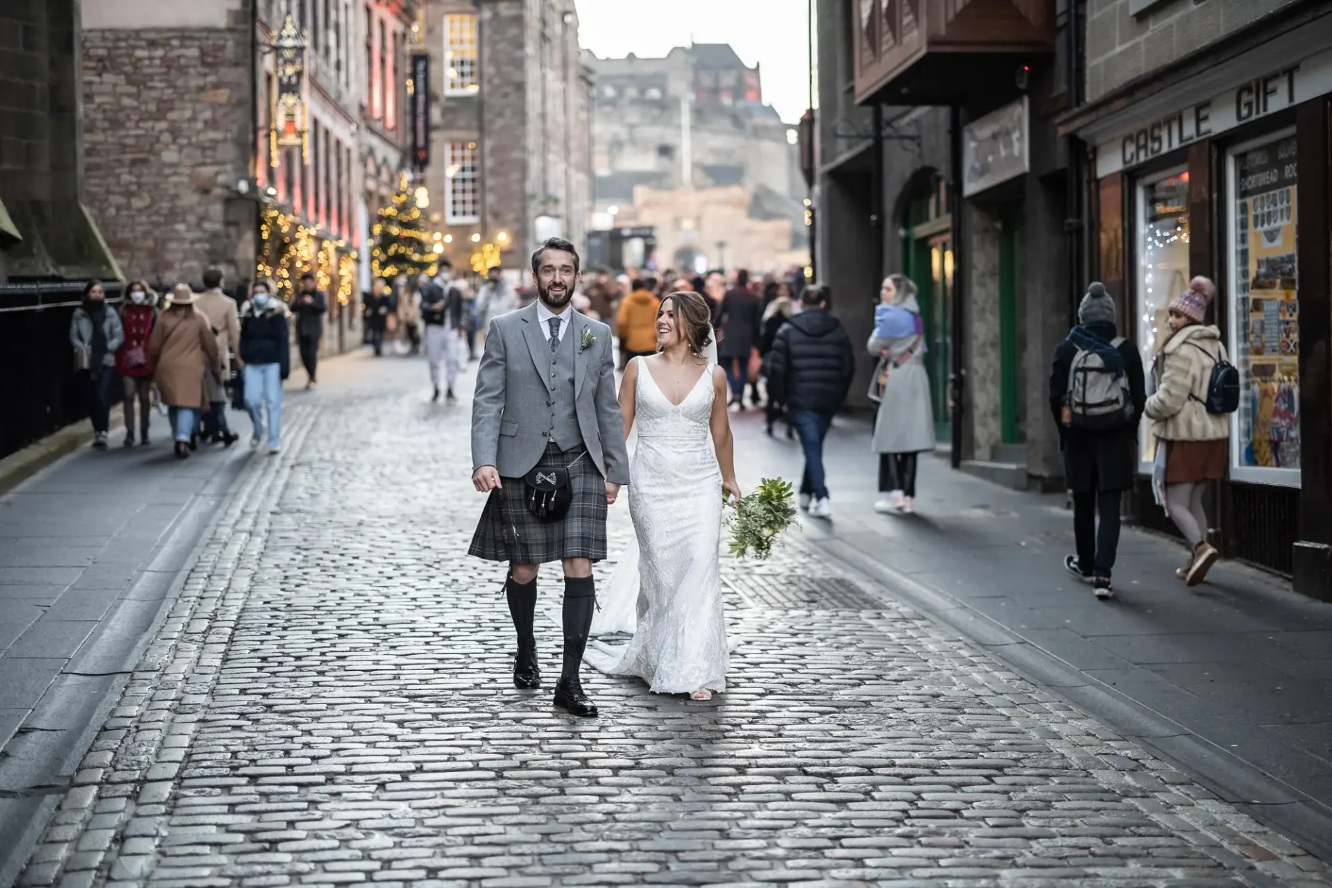A newlywed couple, the groom in a kilt and the bride in a white dress, joyfully walk down a cobblestone street in a busy urban area adorned with festive lights.