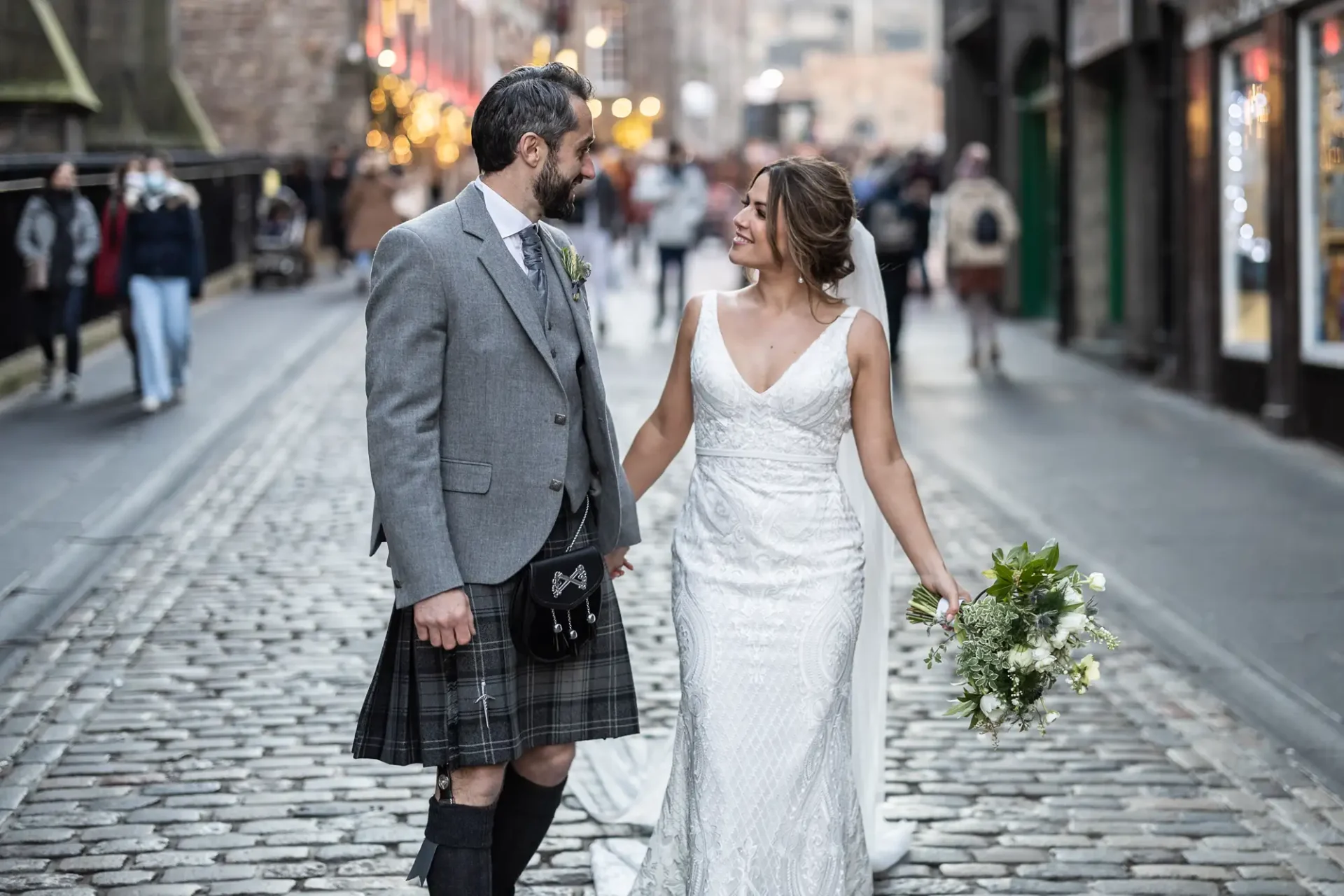 A bride in a white dress and a groom in a kilt sharing a joyful moment as they walk down a cobbled street.