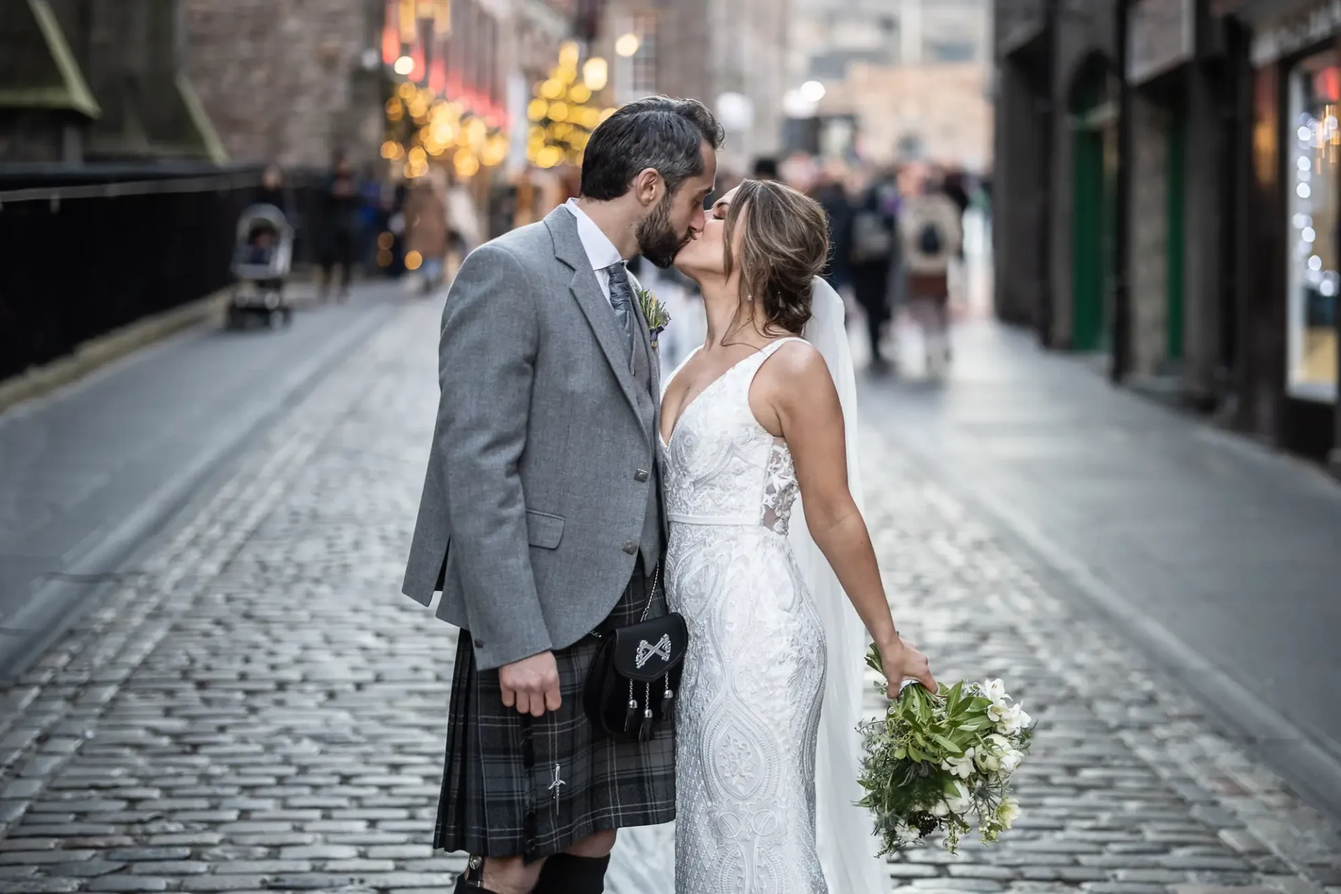 A bride and groom kiss in the middle of a cobblestone street, with the groom wearing a kilt and the bride in a white dress, carrying a bouquet.