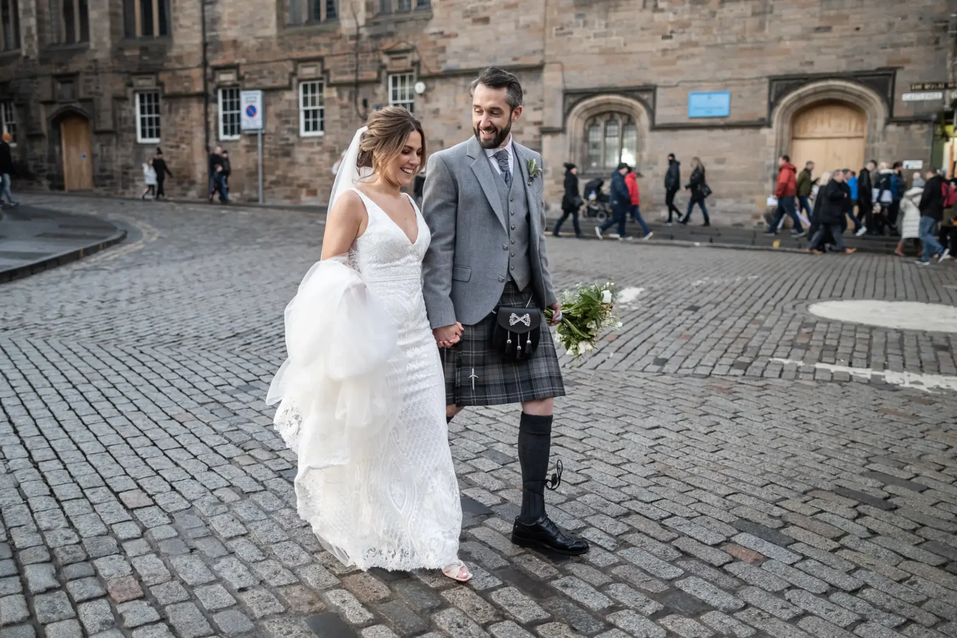 A bride in a white gown and a groom in a kilt holding hands and smiling while walking on a cobblestone street with people in the background.