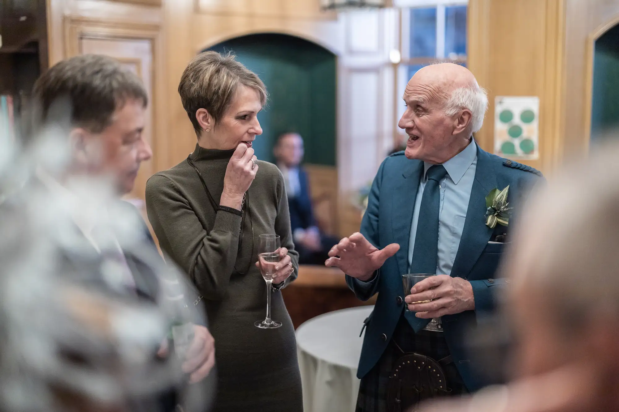 Three people engaged in a lively conversation at a social event, with the older man gesturing animatedly and a woman listening intently while holding a glass of wine.