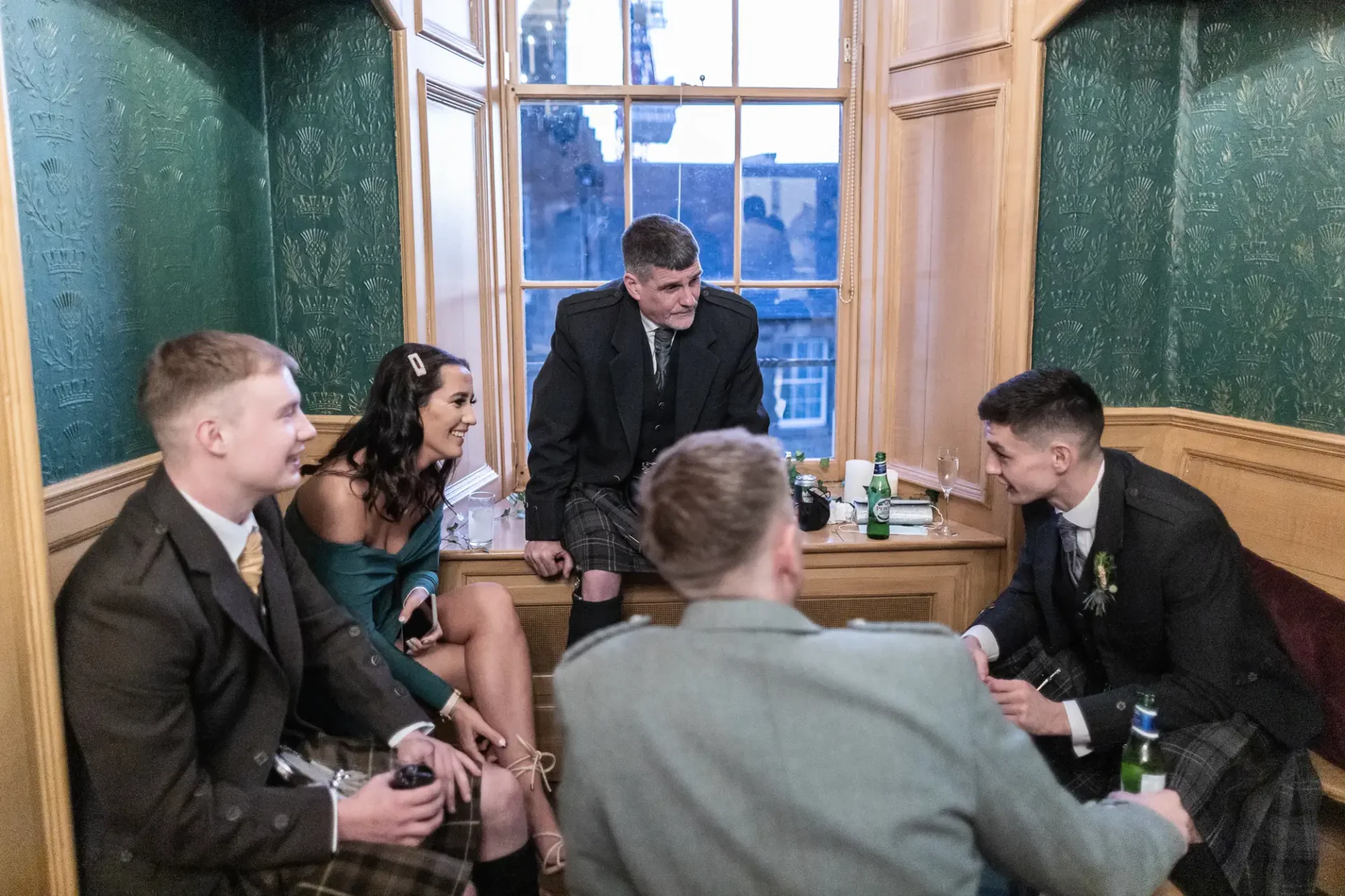 Group of young adults in traditional scottish attire enjoying a lively conversation in an ornate room with green walls and large windows.