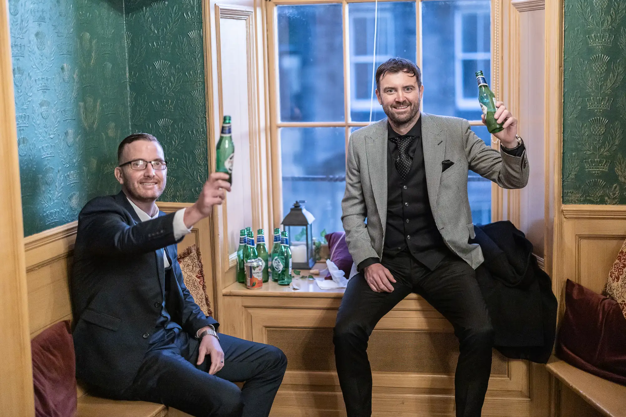 Two men in suits sitting in a classic booth, toasting with bottles of beer, smiling and looking at the camera.