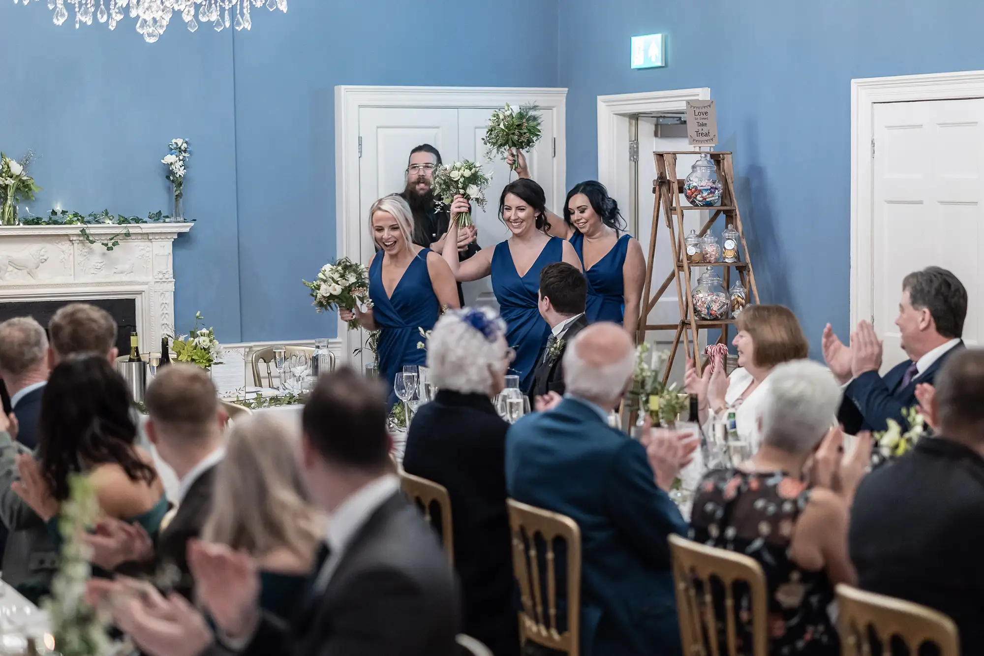 Wedding party entering reception room with guests clapping; two women in blue dresses lead, holding bouquets, followed by a man in a suit.