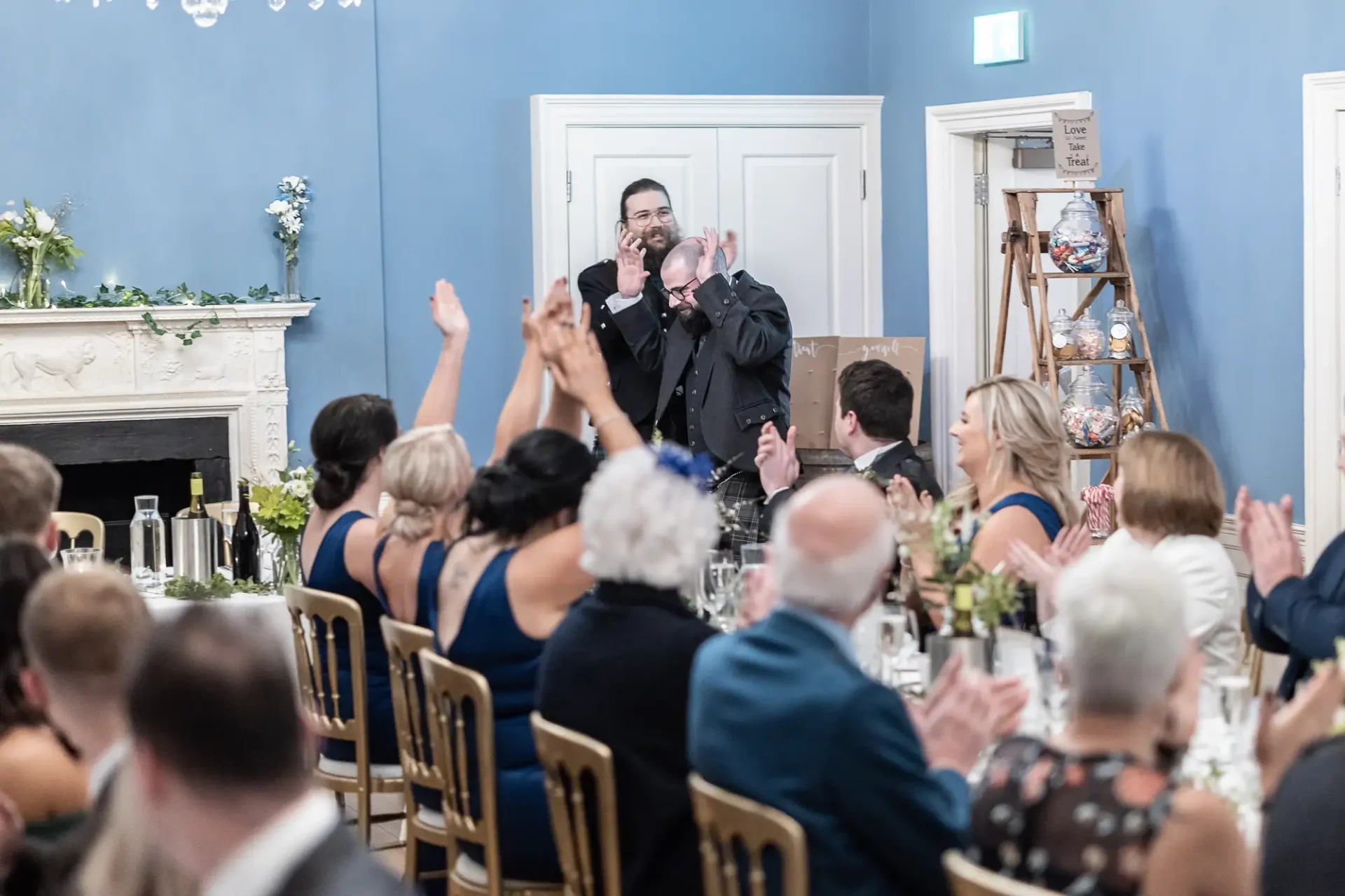 A man giving a speech at a wedding reception, holding a microphone and a glass, with guests seated around tables applauding.