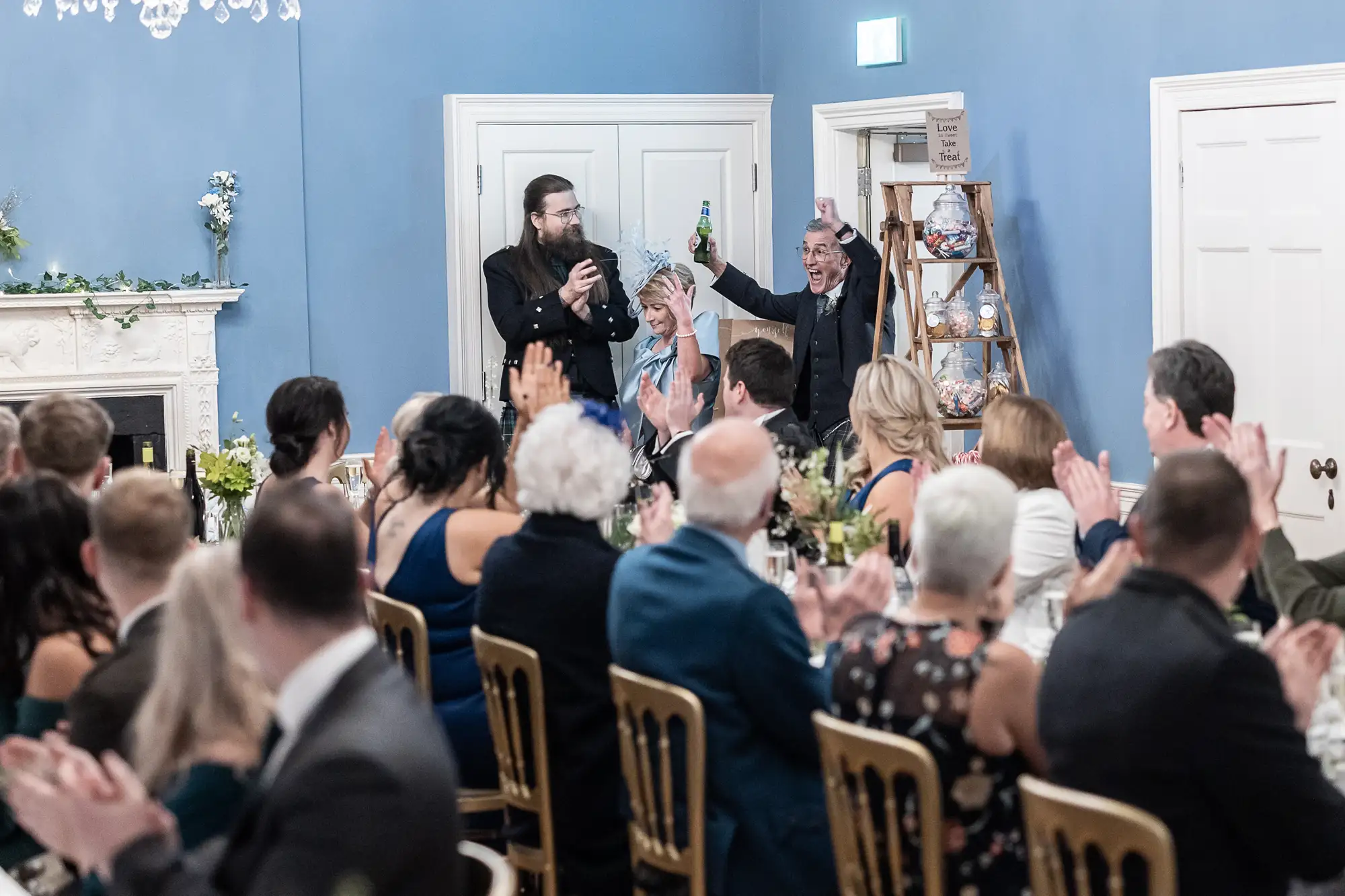 Two men toasting with champagne at a wedding reception in an elegant blue room, surrounded by seated applauding guests.