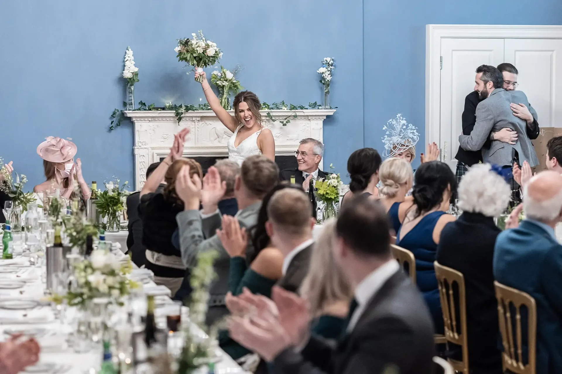 A bride joyfully tossing her bouquet to guests at a wedding reception in an elegantly decorated room with blue walls.
