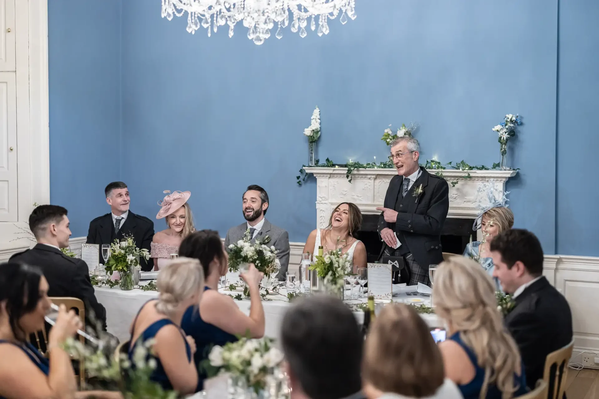 A man gives a speech at a wedding reception in an elegant room with guests seated at decorated tables, laughing and clapping.