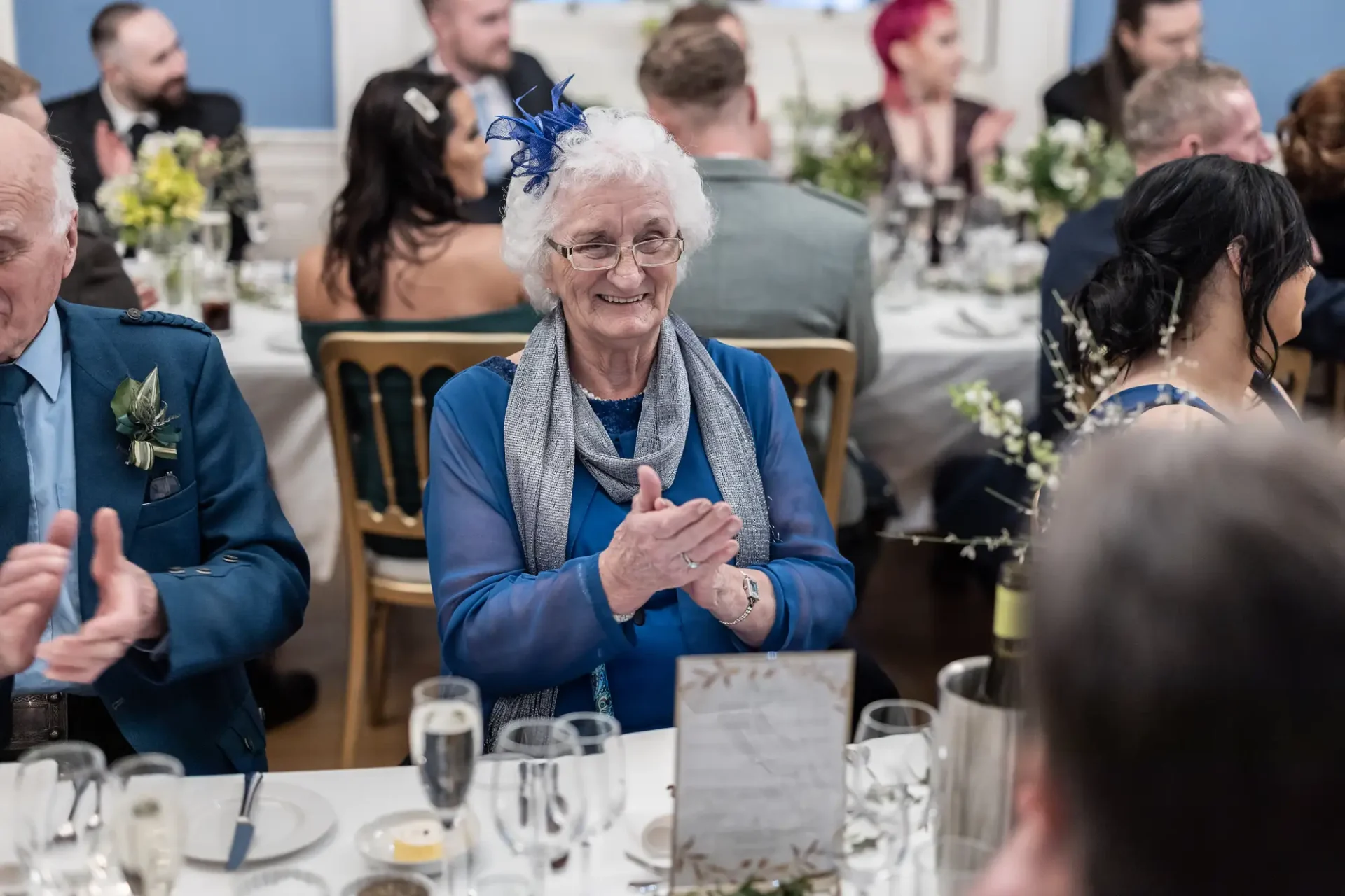 Elderly woman in blue with a feather fascinator smiling and clapping at a wedding reception, surrounded by other guests.