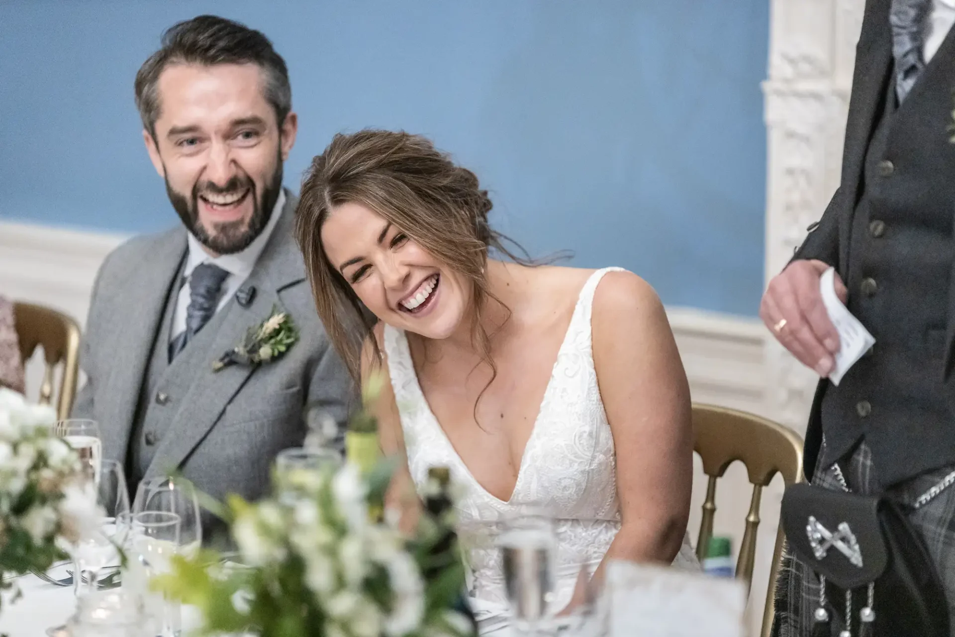 A beaming bride and groom laugh at a wedding reception table, surrounded by elegant dinner settings in a well-lit room.