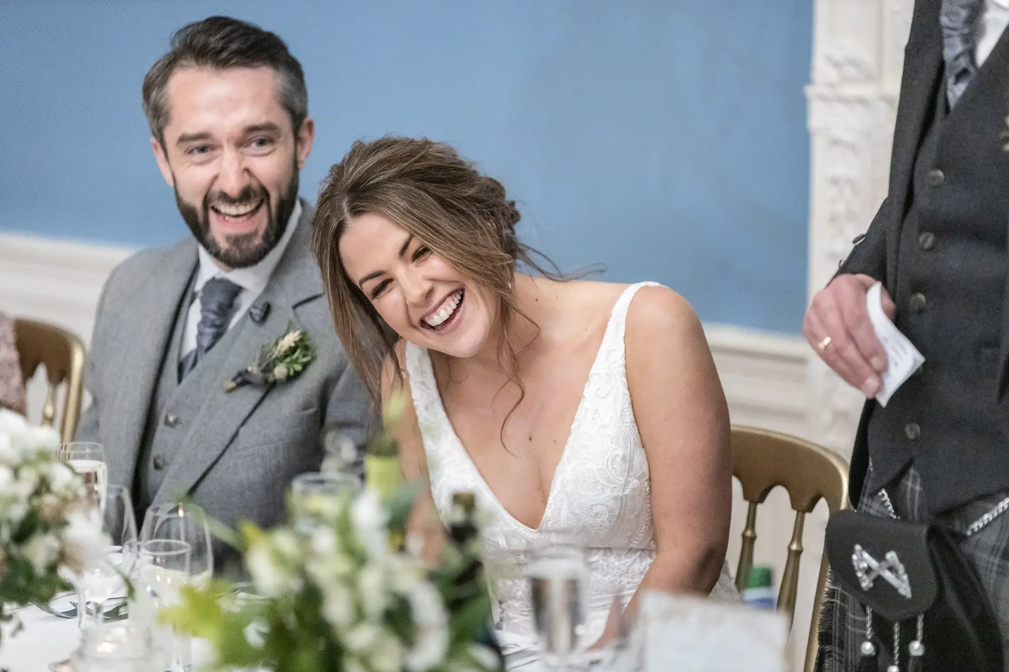 A beaming bride and groom laugh at a wedding reception table, surrounded by elegant dinner settings in a well-lit room.