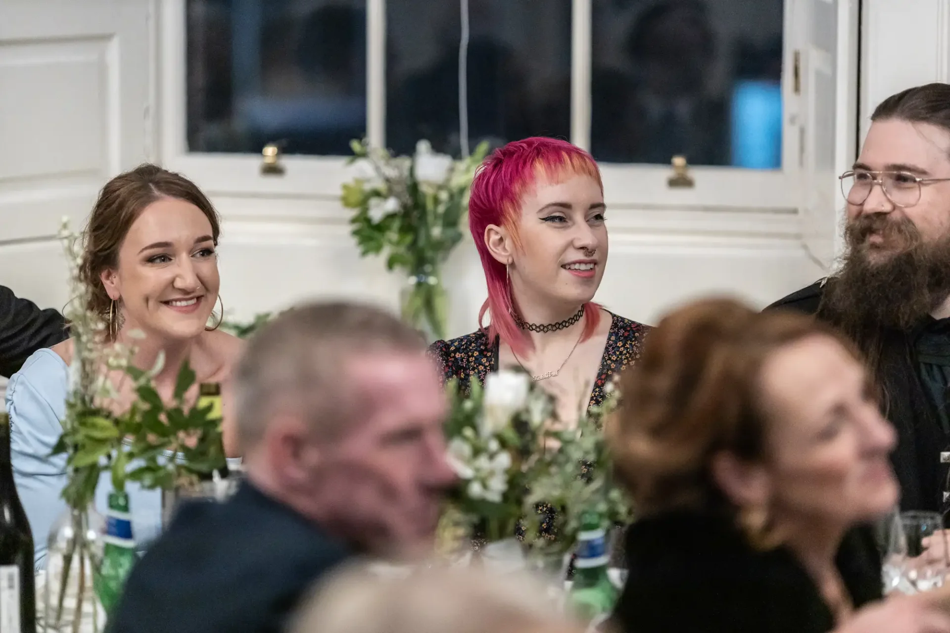 Three people sitting at a dining table during an event, smiling and listening attentively, with a woman in the center sporting bright pink hair.