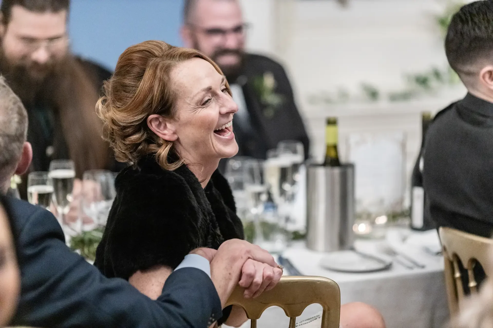 A woman with red hair laughing joyfully at a dinner table during a formal event, surrounded by other guests.