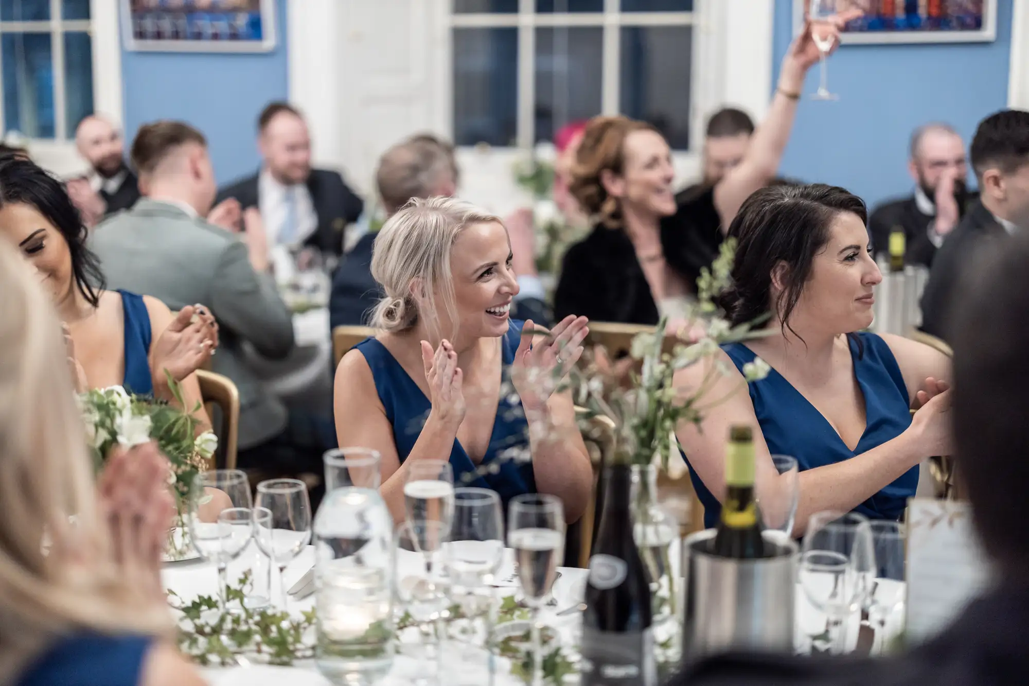 Two women in blue dresses smiling and clapping at a lively banquet with guests engaging in conversations around them.