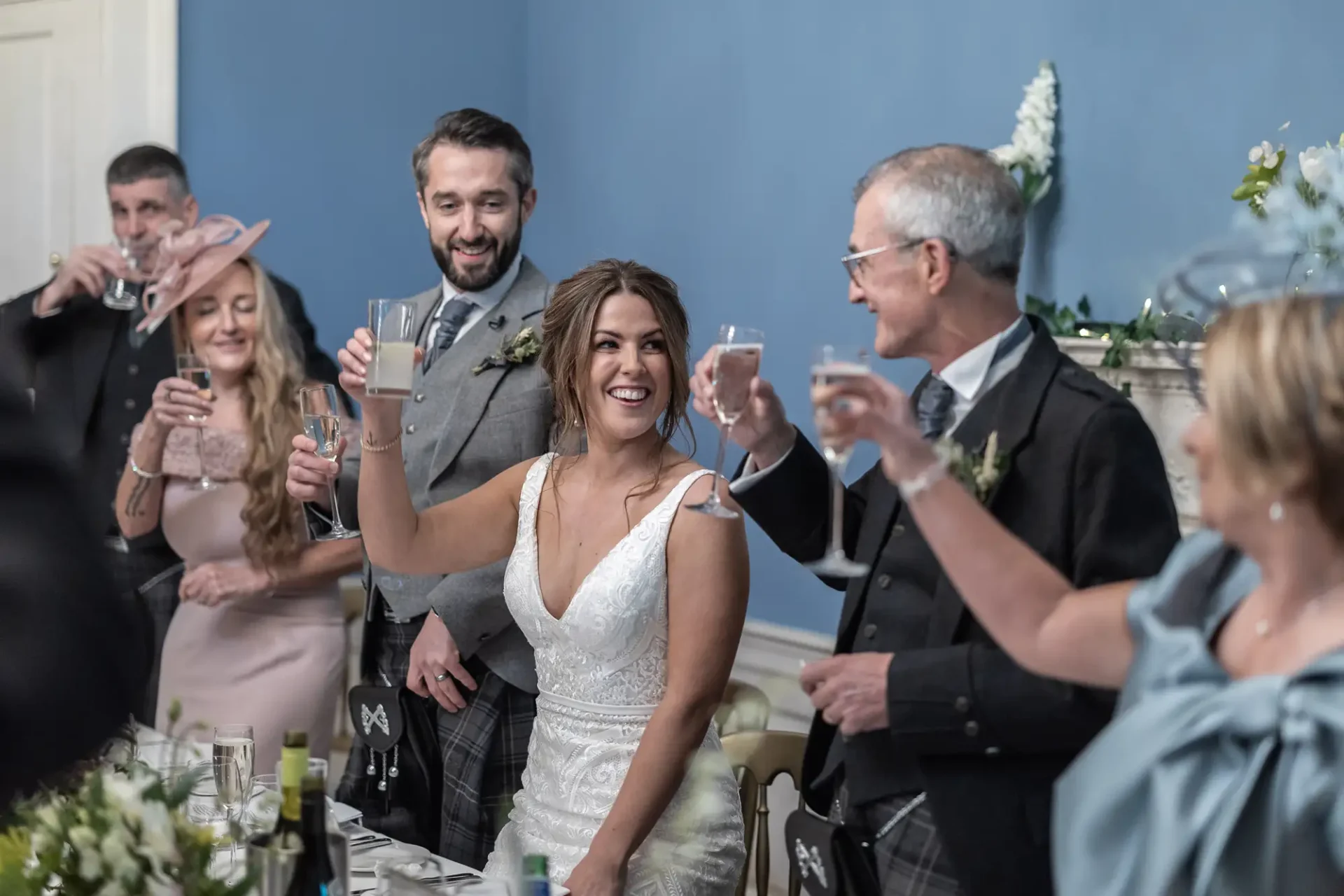 A bride and groom cheerfully toast with guests at their wedding reception, surrounded by smiling friends and family.