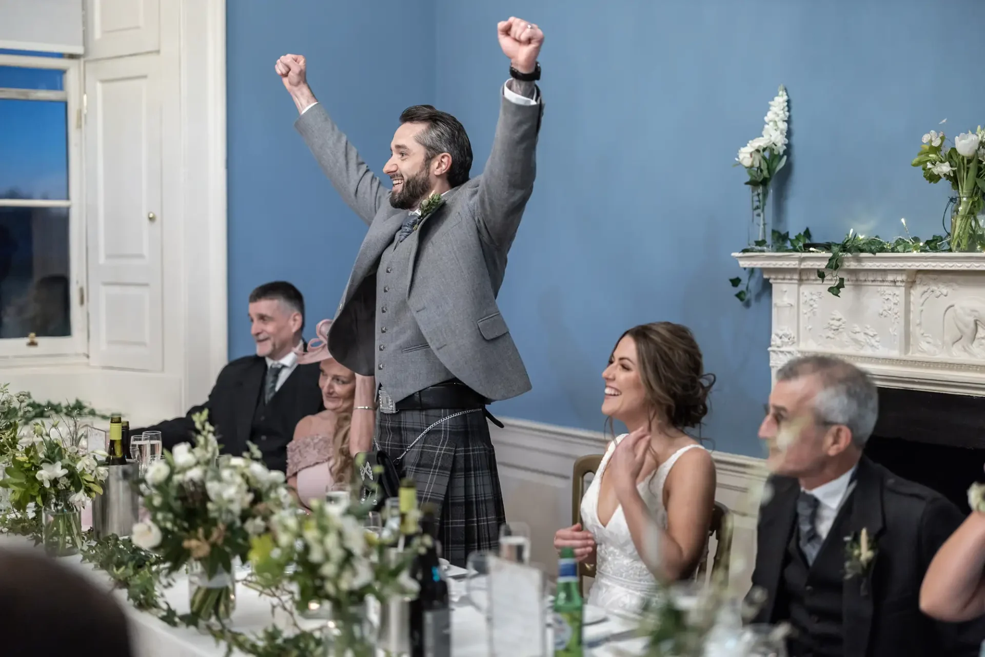 A man in a kilt and vest excitedly raises his arms at a wedding reception, while a smiling woman seated next to him claps, surrounded by other guests.