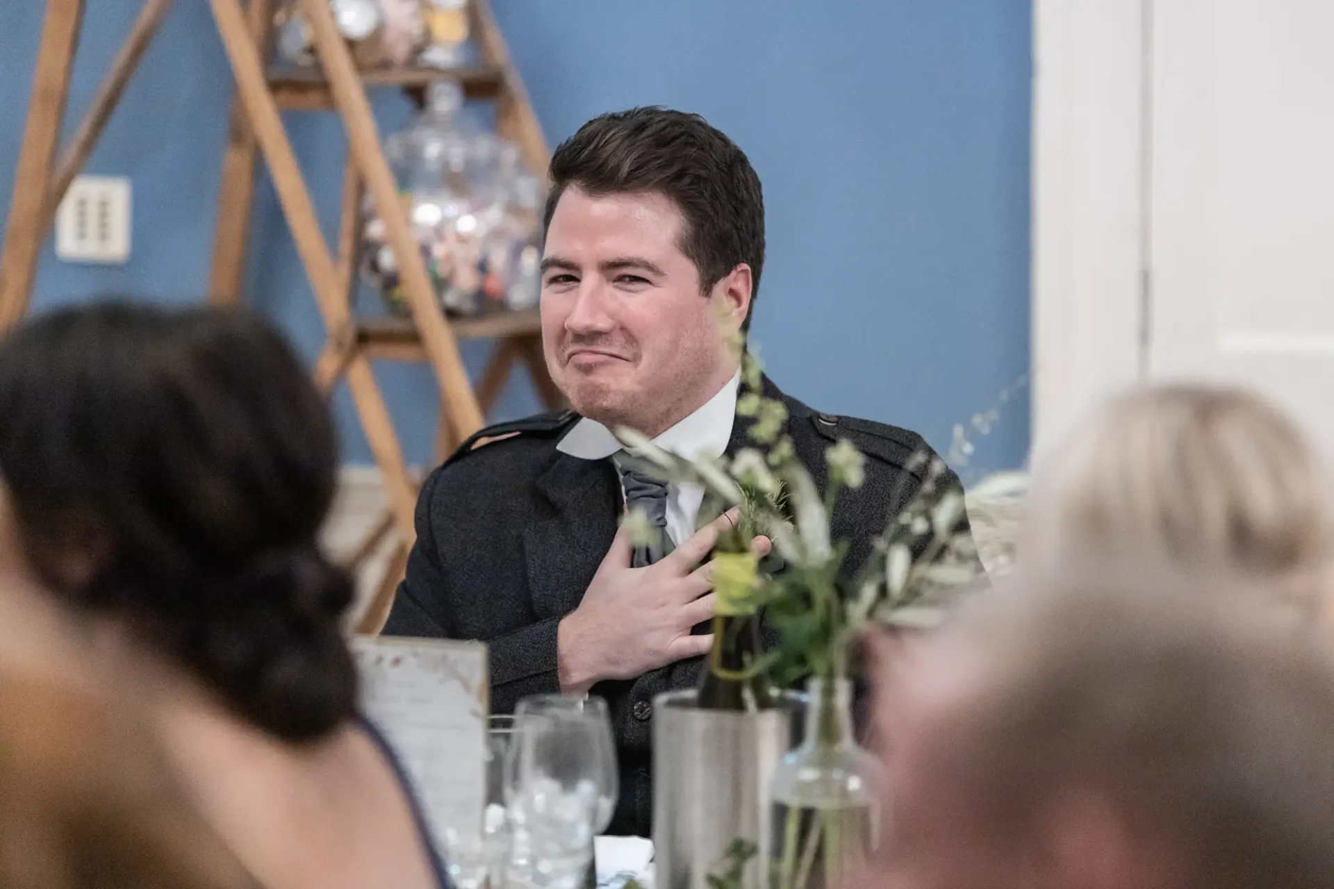 A man in a dark suit making a heartwarming gesture and smiling at a wedding reception table.