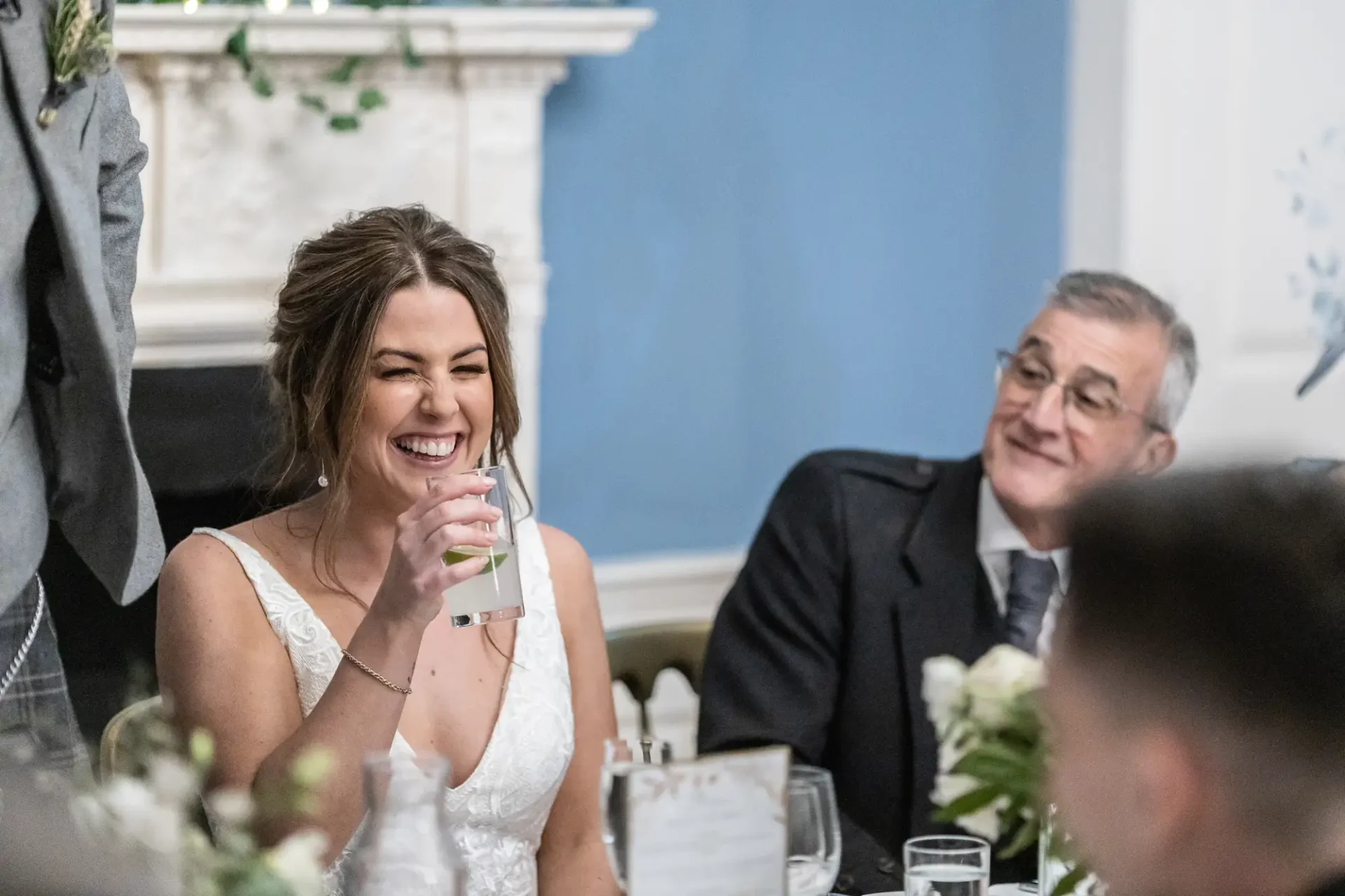 A bride laughing joyfully with a glass in hand at a wedding reception table, a man smiling at her in the background.