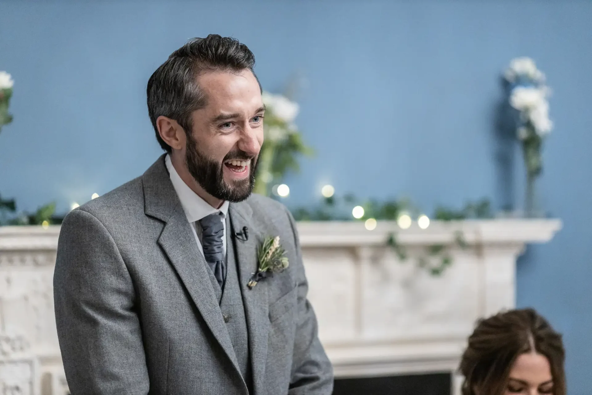 Bearded man in a gray suit with a boutonniere smiling at a wedding, with a woman and decorated mantelpiece in the background.
