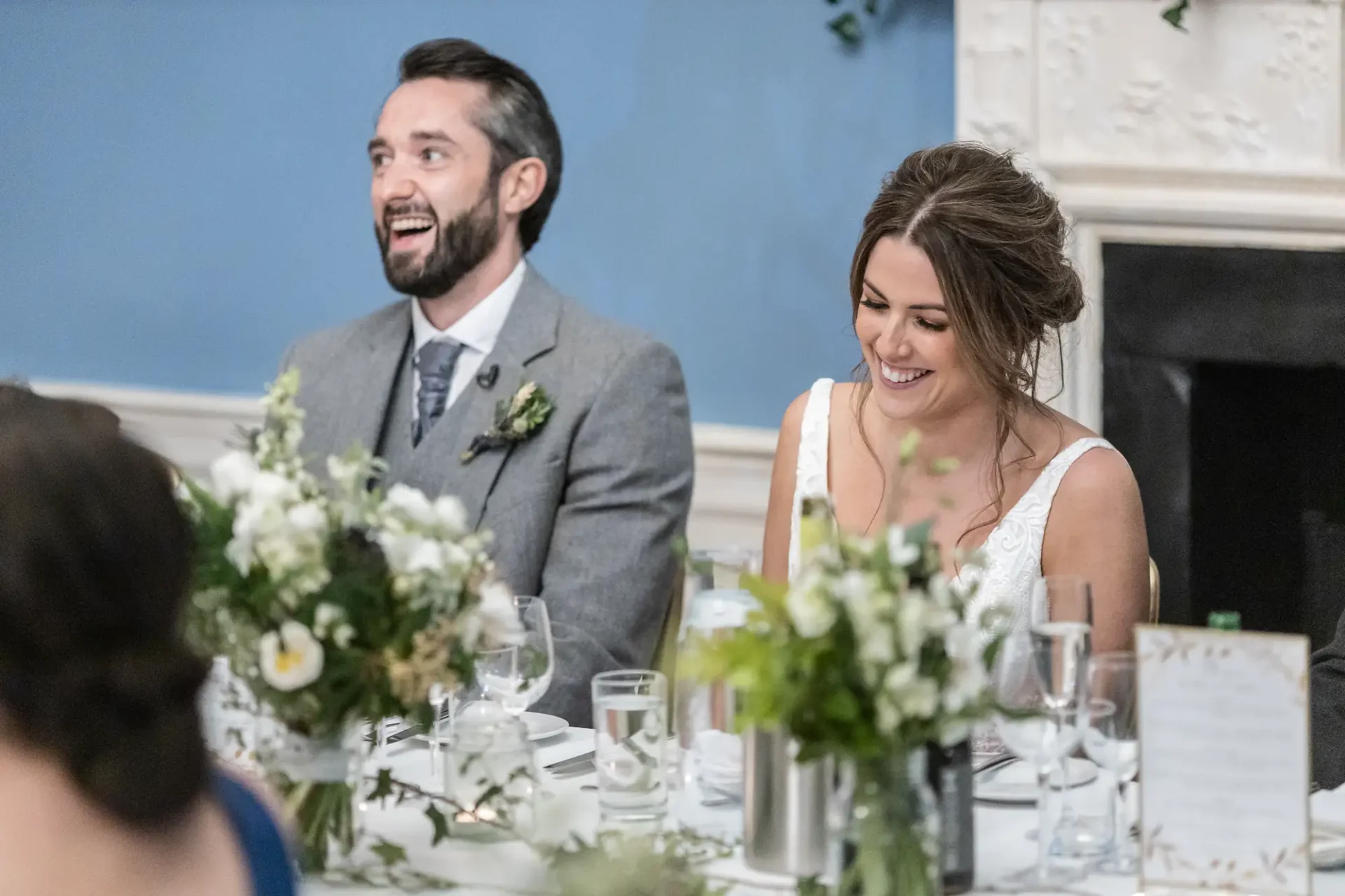 A bride and groom laugh joyfully at a wedding reception table, surrounded by elegant floral arrangements.