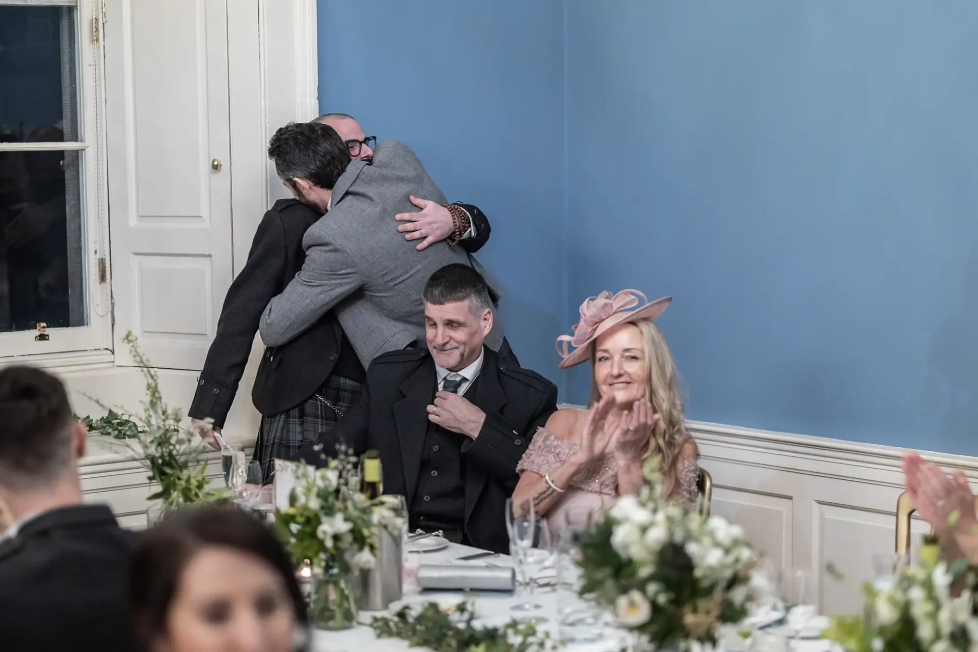 Two men embracing at a wedding reception while a seated man and woman look on, smiling, in a room with light blue walls and floral decorations.