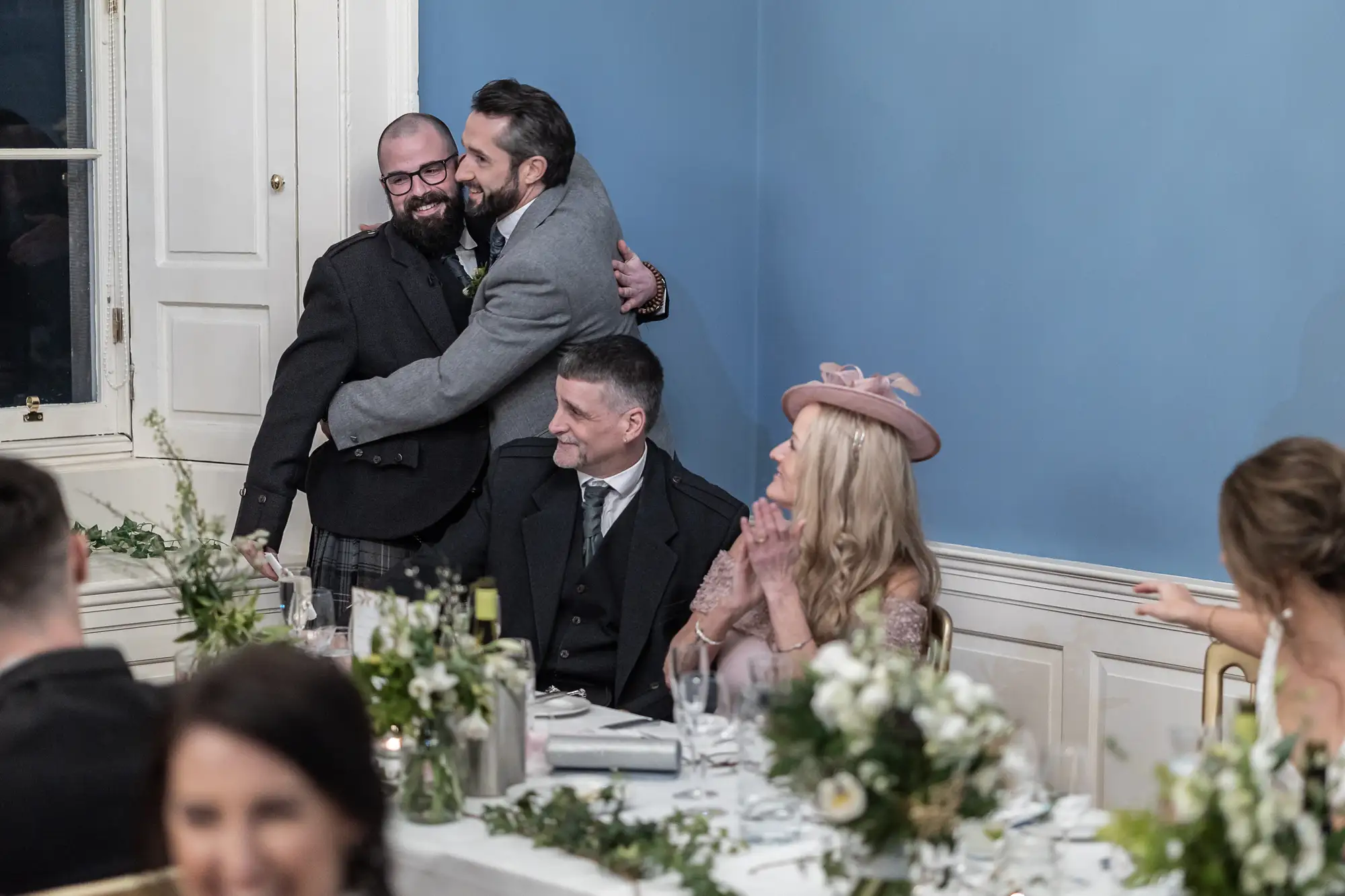 Two men embracing happily at a wedding reception, with guests seated at tables around them, all in a room with blue walls and elegant decor.