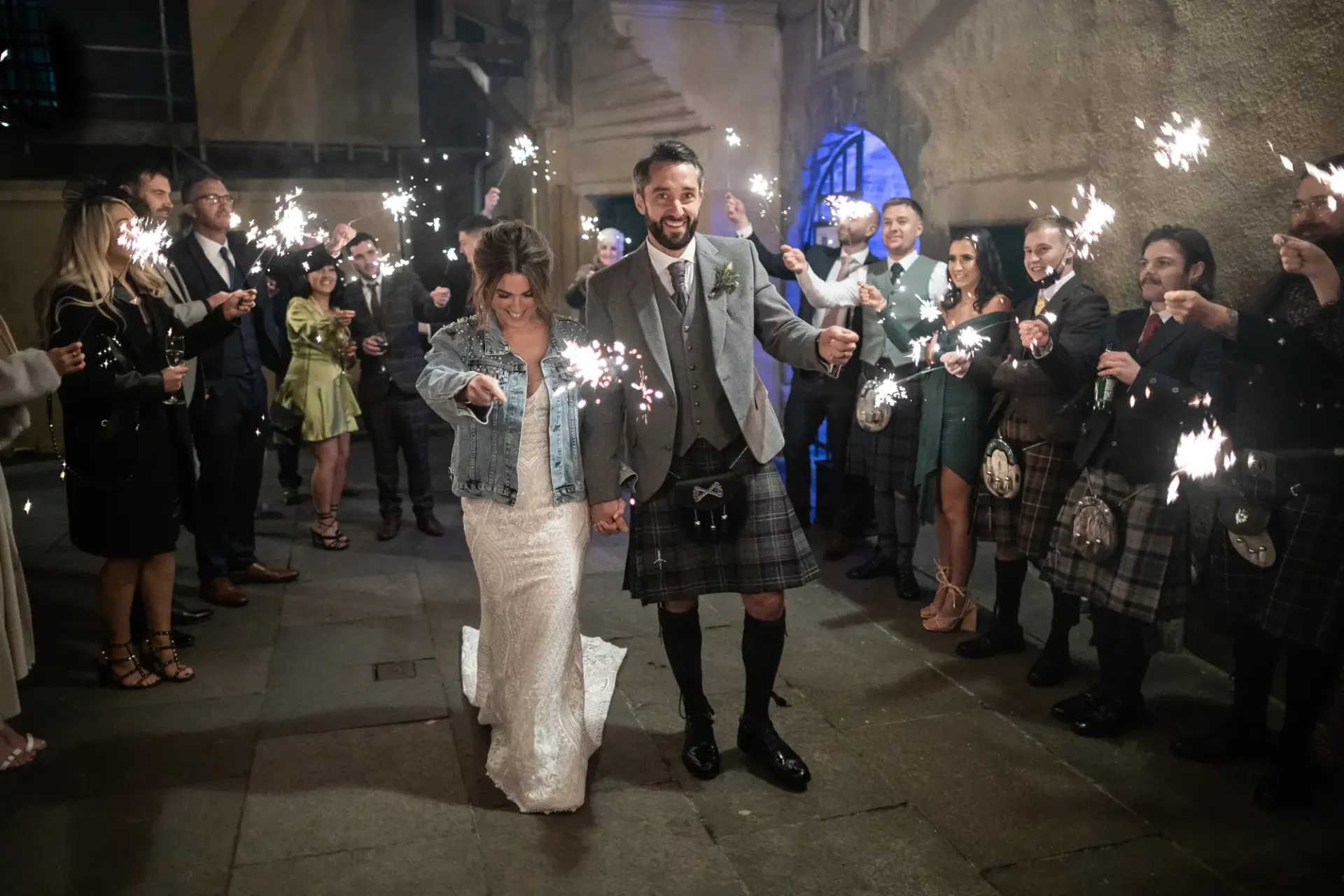 A bride and groom walk through a tunnel of guests holding sparklers, the groom in a kilt and the bride in a denim jacket.