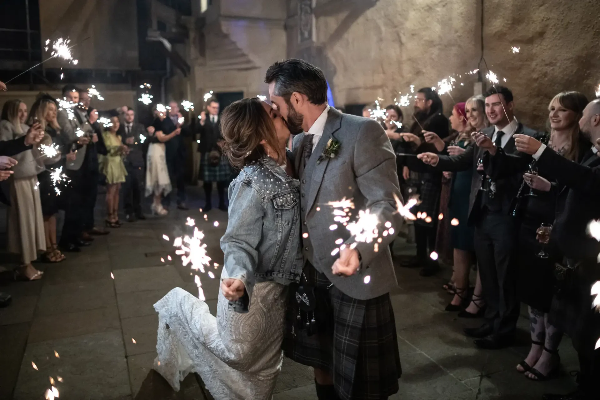 A couple kissing during a sparkler send-off at night, surrounded by guests in a festive setting.