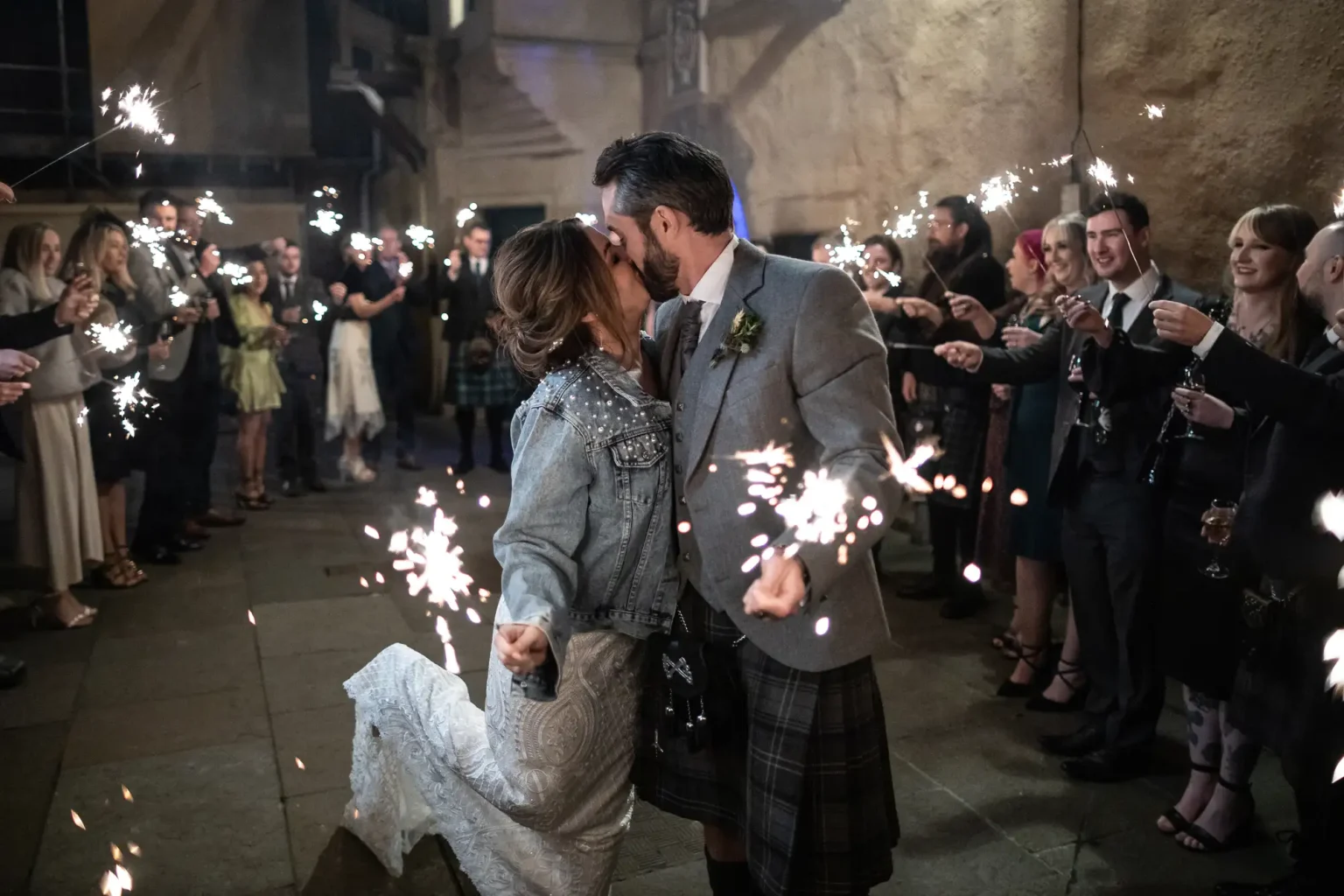 A couple kisses while surrounded by guests holding sparklers at a nighttime wedding celebration.