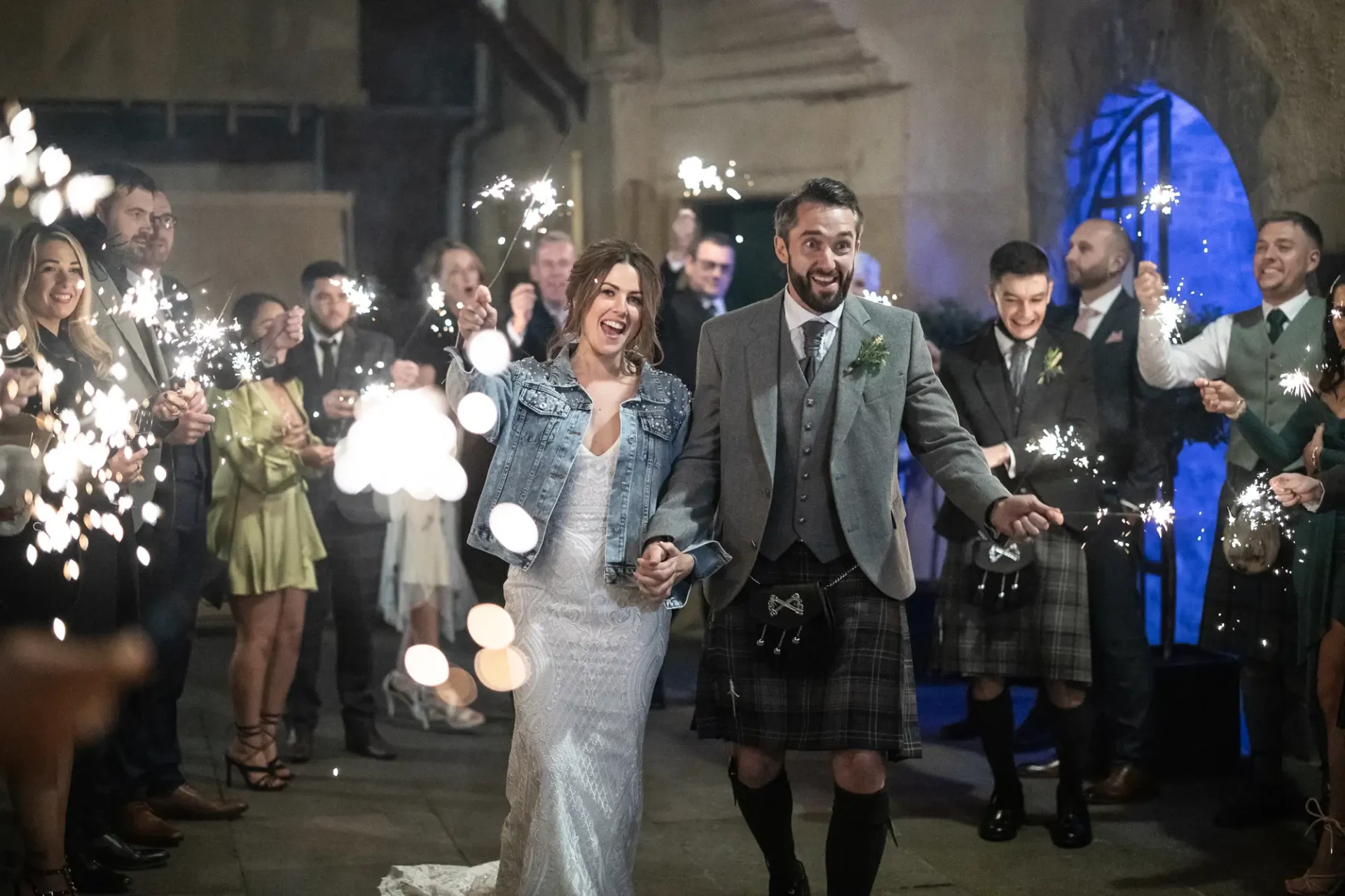 A joyful bride and groom walk hand-in-hand through a sparkler send-off, surrounded by smiling guests at nighttime. the groom is dressed in a kilt, and the bride wears a white dress and denim jacket.