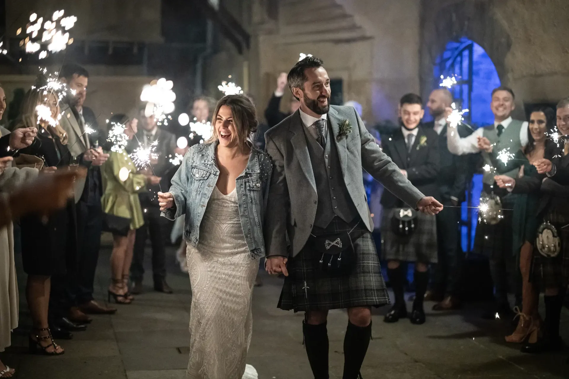A joyful bride and groom walk through a sparkler send-off at night, with guests lined up on both sides, the groom in a kilt and the bride in a white dress and denim jacket.