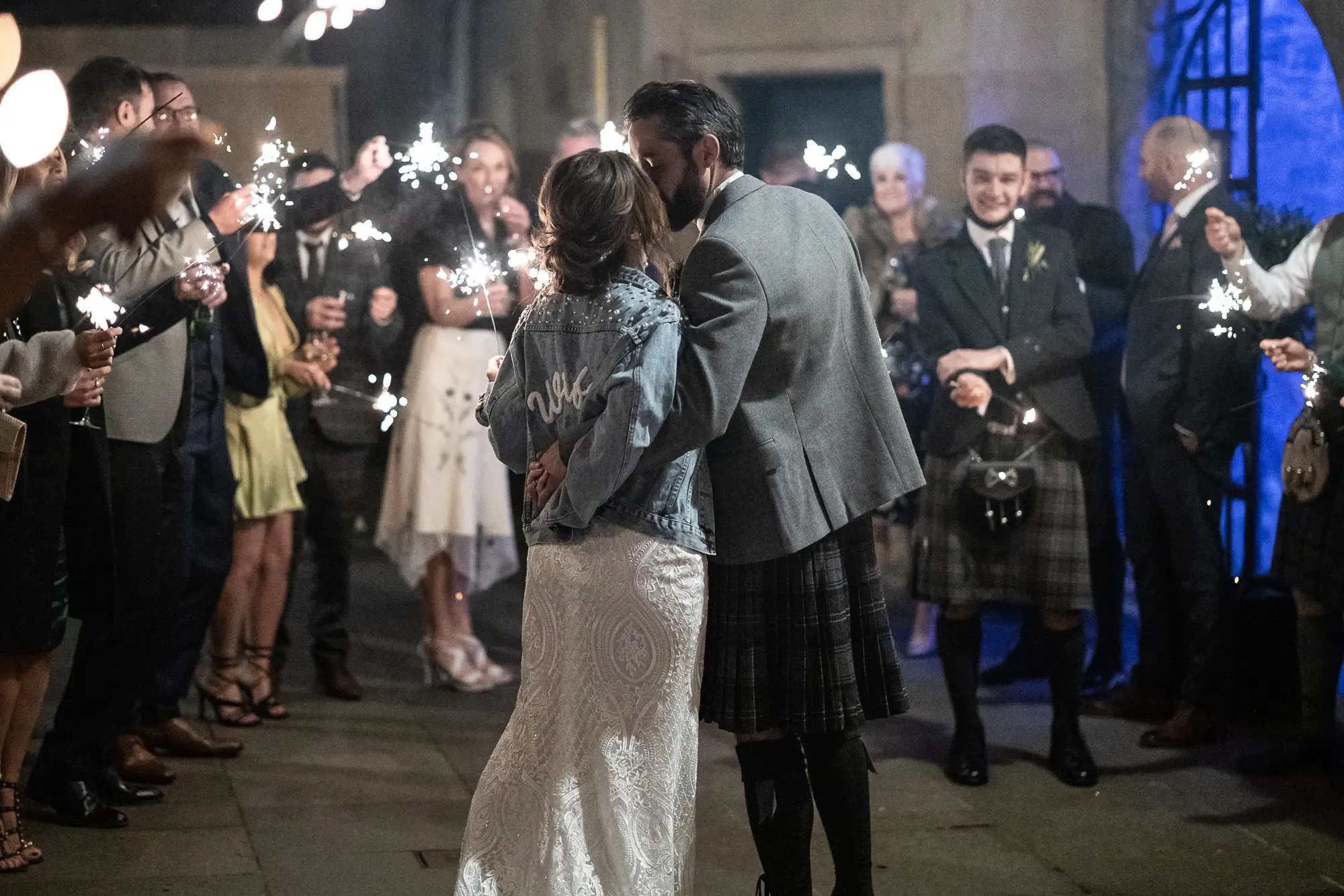 A bride and groom share a kiss surrounded by guests holding sparklers at a night-time wedding celebration.