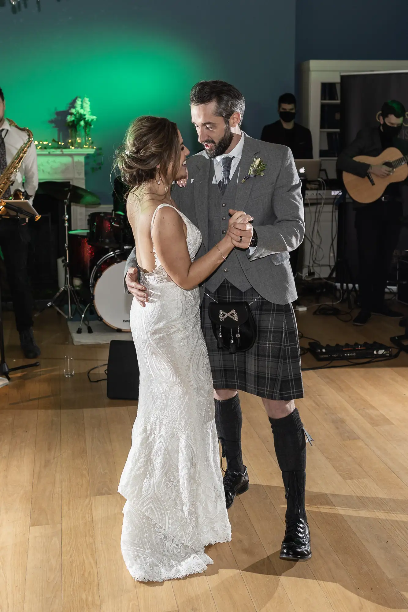 A bride and groom share a dance, the groom in a kilt and sporran, with a live band in the background, at their wedding reception.