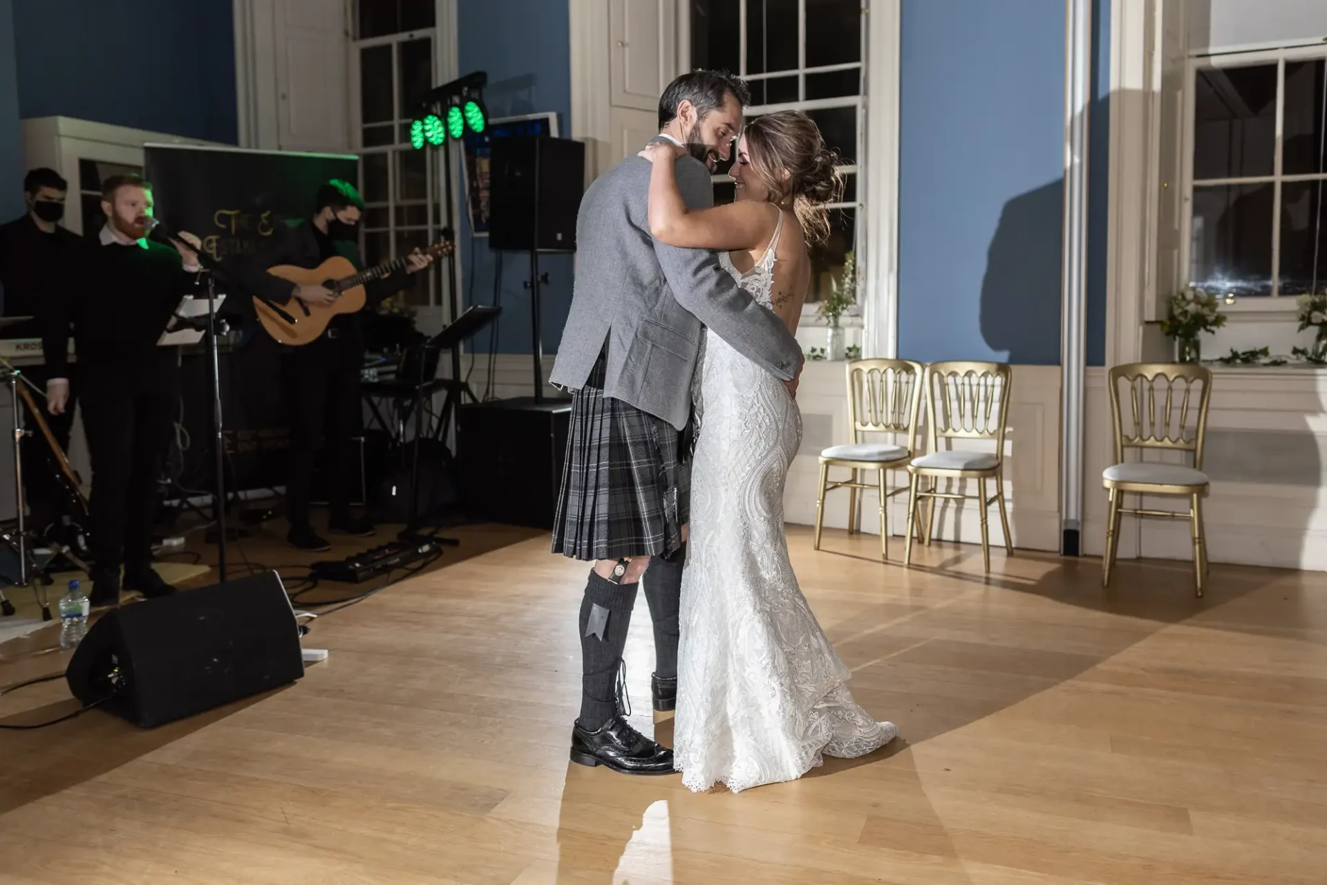A couple in wedding attire, the groom in a kilt and the bride in a lace gown, share a dance in a ballroom with a live band in the background.