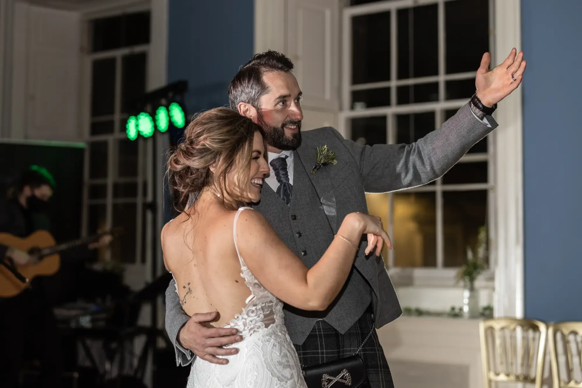 A bride and groom dancing joyfully at their wedding reception, the groom gesturing with one arm as a band plays in the background.