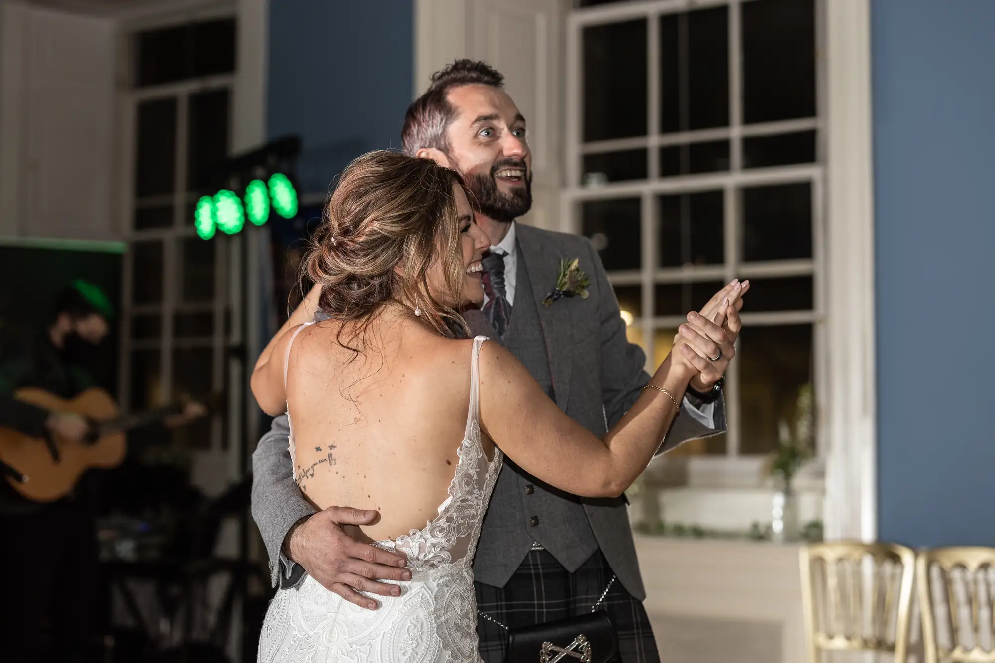 A newlywed couple dances closely, the man in a kilt and the woman in a white dress, both smiling joyously in a ballroom with a musician in the background.