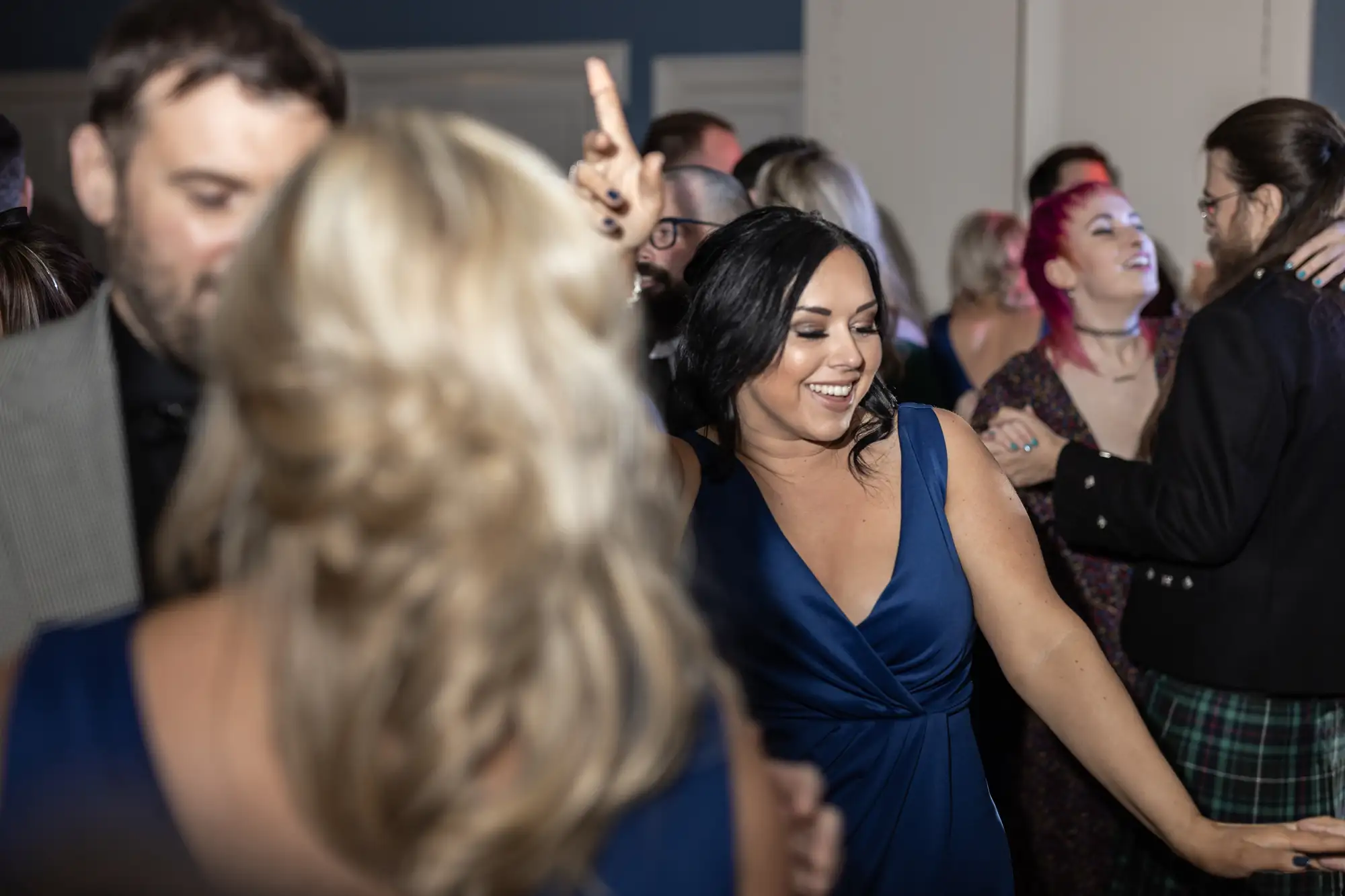 A joyful woman in a blue dress laughs while interacting with people at a crowded indoor event.