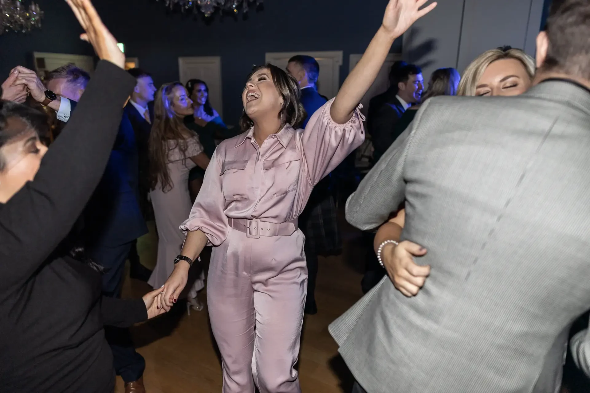 A woman in a pink jumpsuit laughing joyfully while dancing at a crowded party.
