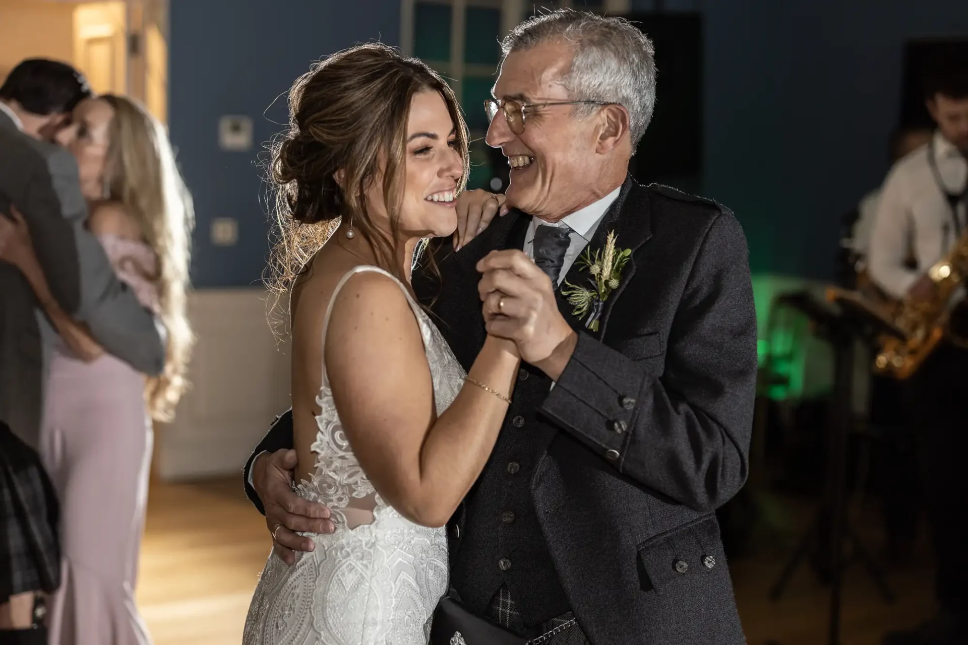 A bride and her father share a joyful dance at her wedding reception, both smiling widely in a warmly-lit hall with guests and a live band in the background.