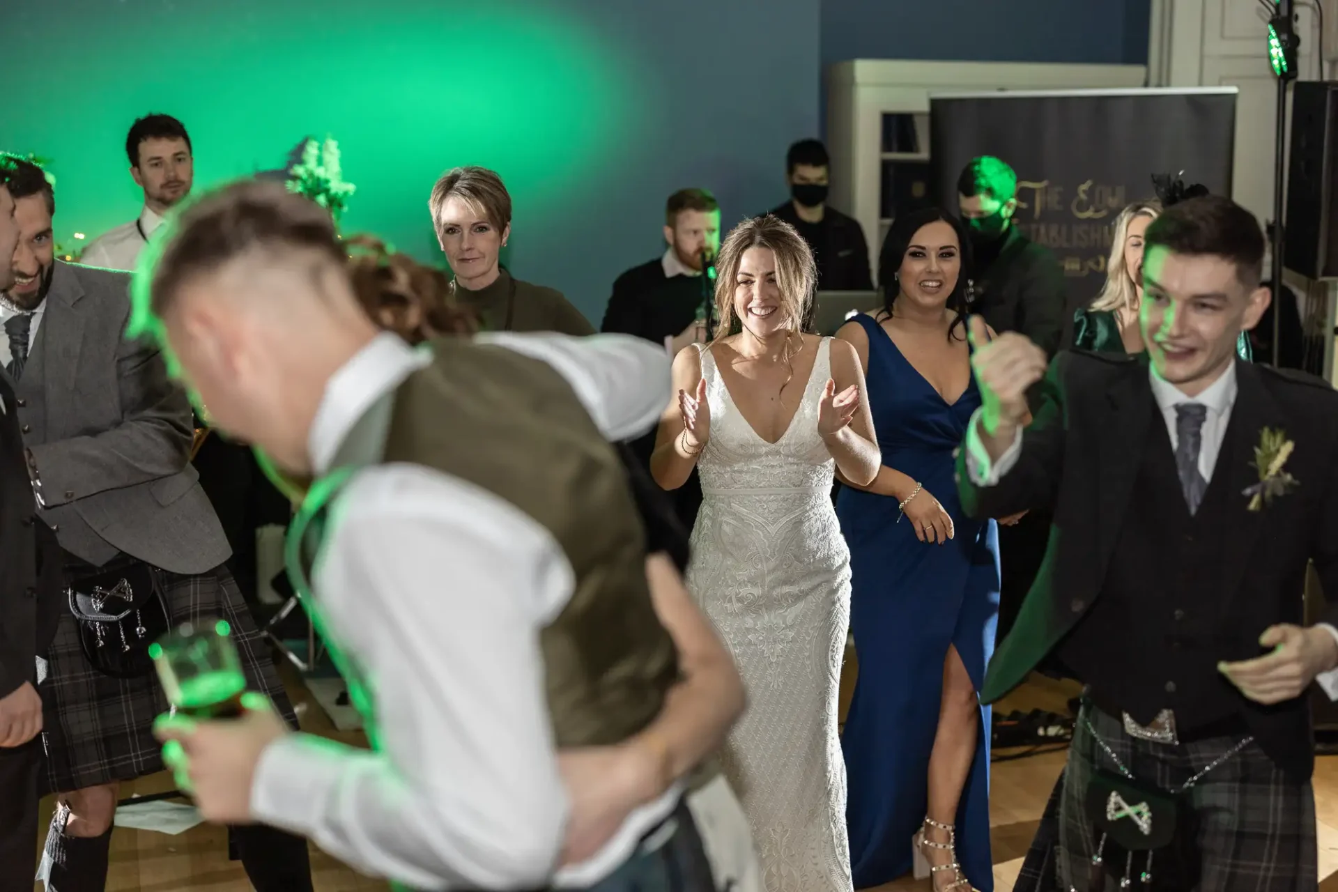 A bride laughs joyfully while dancing with guests at a wedding reception. some men are dressed in kilts; a green light adds ambiance in the background.